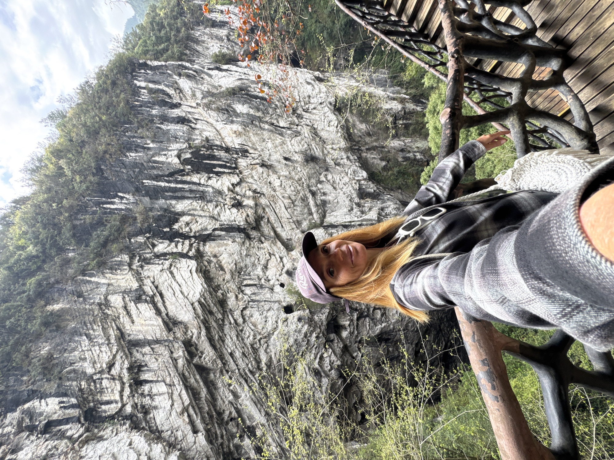 A blonde woman wearing a plaid shirt and a baseball cap is taking a selfie on a wooden structure in Zhuoshui, China. The structure has a railing made of natural-looking branches painted black. In the background, a large, rocky cliff face dominates the scene, with some greenery clinging to its surface. New growth, likely from spring, is visible near the woman and the structure. The sky is overcast.