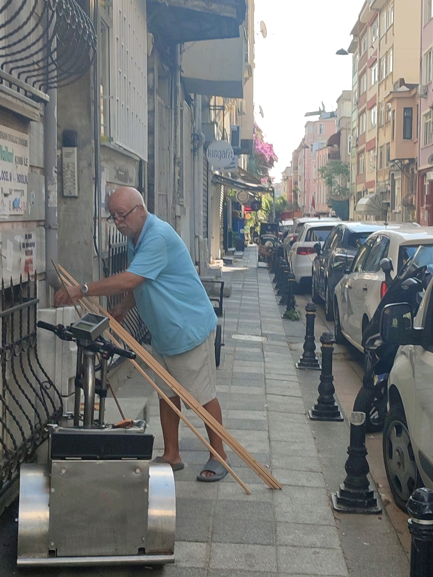 The image depicts a street scene in Kadıköy, Turkey. In the foreground, a bald, middle-aged man wearing glasses, a light blue polo shirt, and khaki shorts is interacting with a metal cart. He is holding several long, slender wooden poles. The cart appears to be a portable cleaning or maintenance device, with a metallic casing and wheels. The man is standing on a paved sidewalk. Behind him, a black wrought-iron fence runs along the side of a building, which has posters and signage on its exterior. In the background, a narrow street is lined with multi-story buildings with shops on the ground floor. Several cars are parked along the street, and decorative bollards line the sidewalk. The overall impression is of a typical urban environment in Turkey.