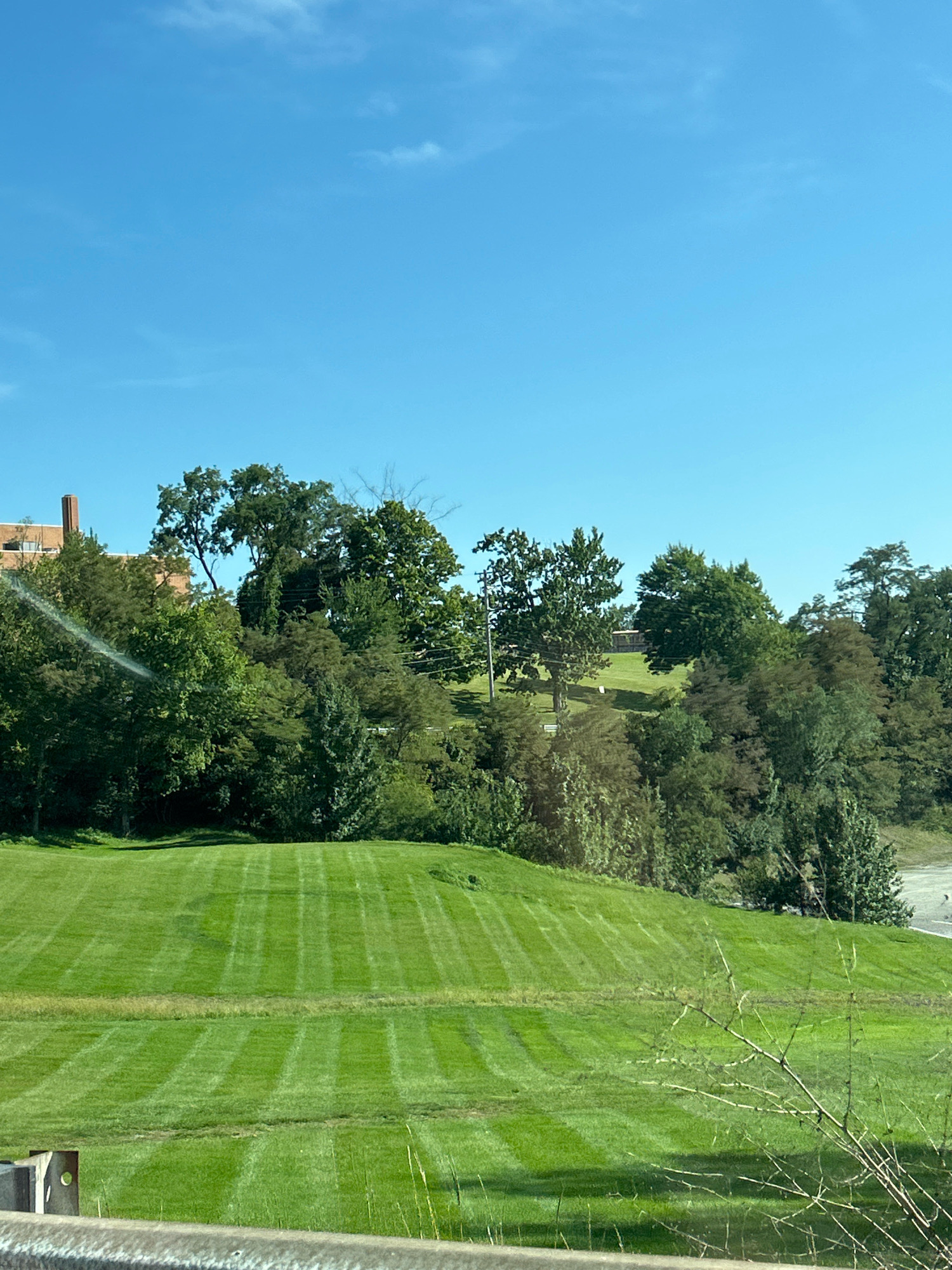 The image depicts a landscape scene under a clear blue sky with a few wispy clouds. In the foreground, a neatly mowed green lawn displays distinct striped patterns from a lawnmower, suggesting recent maintenance. A low barrier, possibly a guardrail, is visible in the bottom left corner of the frame. Beyond the lawn, a gentle slope is covered in more of the same striped grass, leading up to a dense collection of trees.
To the left of the trees, a brick building with a prominent chimney is partially visible, hinting at an institutional or residential structure. The trees are a mix of deciduous and possibly evergreen types, varying in height and density, with some exhibiting a rich green foliage. Power lines and poles are subtly integrated into the background, suggesting the presence of infrastructure. The overall impression is one of a well-maintained park-like environment, potentially part of a larger estate, campus, or public area within Robinson Township, United States of America.