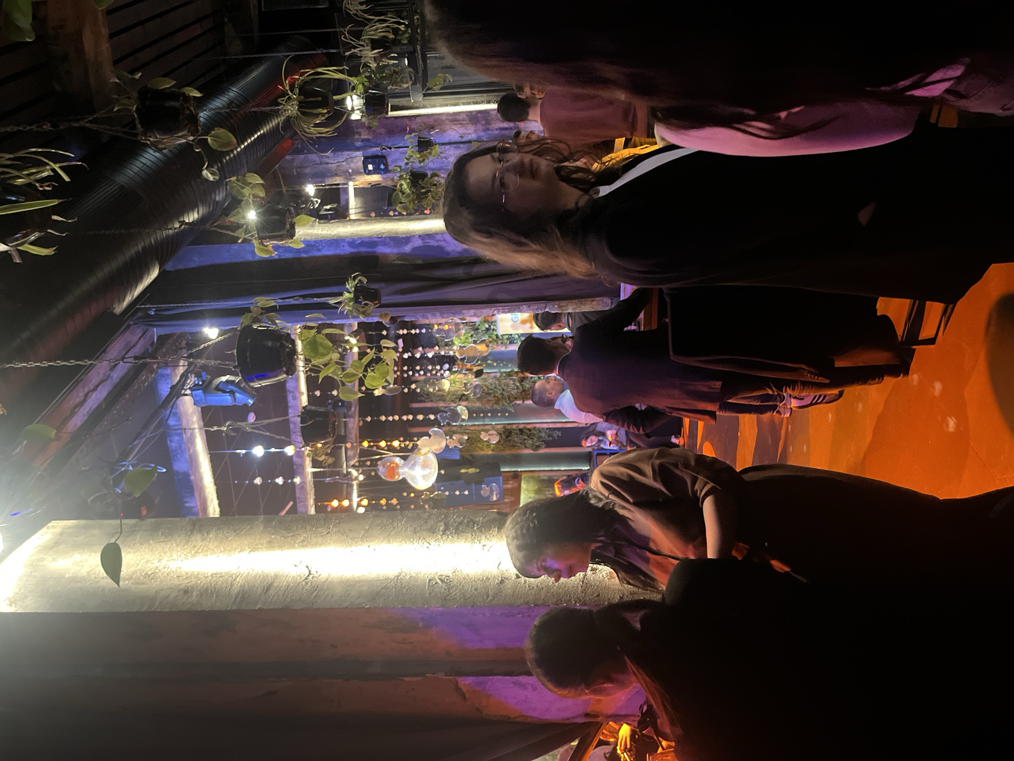 This media file depicts an indoor social gathering, likely in a bar, restaurant, or event venue in Sofia, Bulgaria. The scene is characterized by its dim, atmospheric lighting and eclectic decor.
Several people are present, socializing in various parts of the room. In the foreground, two individuals, mostly silhouetted, are seen leaning over something, possibly a table or counter. In the mid-ground, a woman with long hair and glasses looks towards the camera, while other patrons are visible standing and sitting, engaged in conversation.
The lighting plays a crucial role in setting the mood, with strong blue and purple hues washing over the walls and ceiling, complemented by warm orange light on the floor and other surfaces. Numerous decorative light sources are visible, including strings of small bulbs, larger hanging glass bulbs with visible filaments, and bright white accent lights along a concrete ledge.
The venue features an industrial-chic design, incorporating large, dark exposed ductwork or pipes along the ceiling. Contrasting with this are numerous potted plants suspended from the ceiling by chains, creating a lush, 'urban jungle' aesthetic. Exposed concrete or plaster walls are also visible.
The overall impression is of an evening or nighttime event, given the reliance on artificial and mood lighting. People appear relaxed and engaged in conversation. No discernible text is visible within the image.