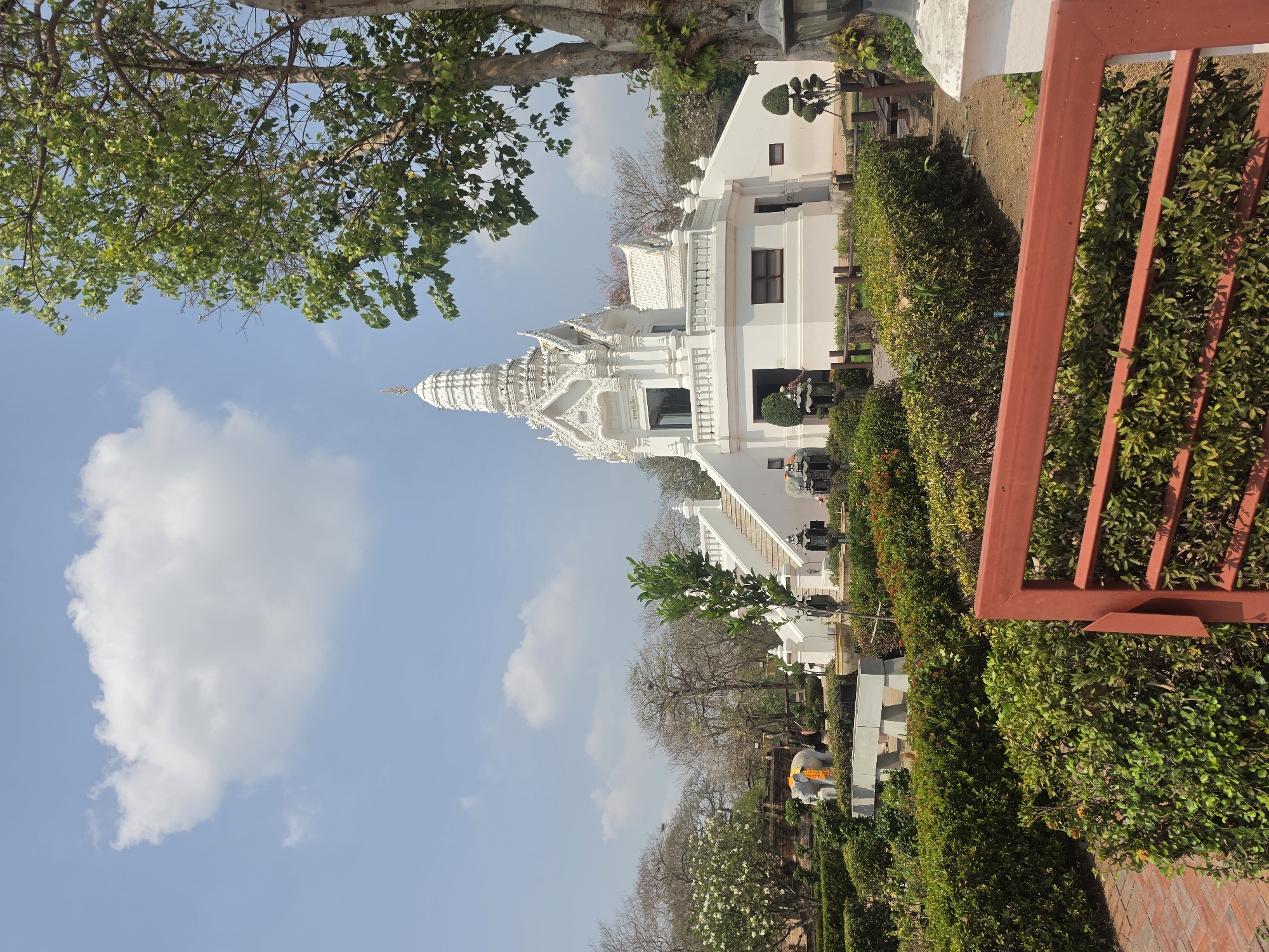 The image depicts a white temple complex in the Historic City of Ayutthaya, Thailand. The temple is multi-tiered with ornate detailing. It is flanked by landscaped gardens with trimmed hedges. A tree with green leaves overhangs the left side of the frame. In the foreground, a red metal fence is visible. The sky is blue with scattered white clouds.