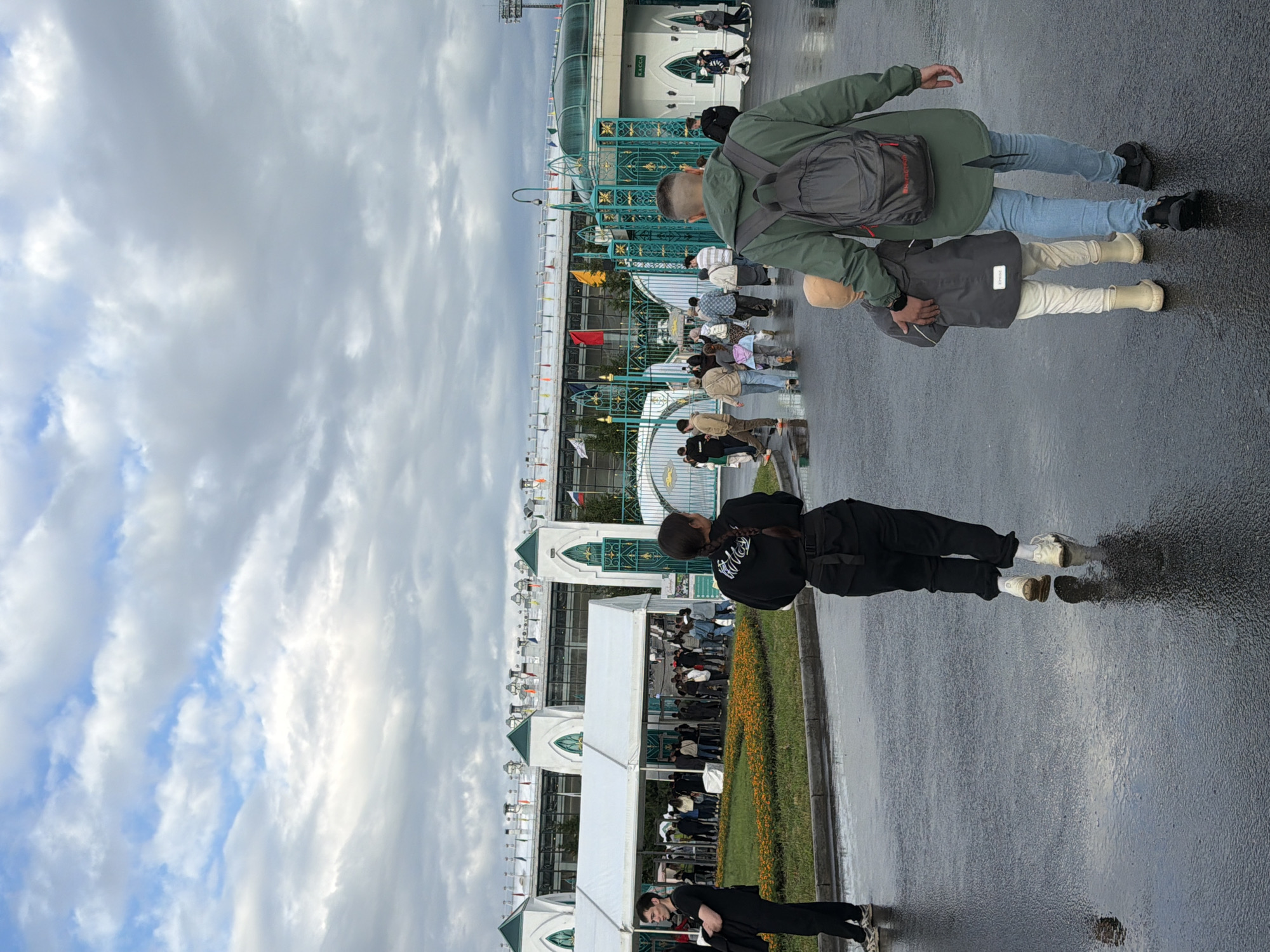 The image depicts an outdoor scene with a group of people walking on a wet paved surface, possibly after rain. In the foreground, two individuals are walking away from the camera. One is a person wearing black clothing, with their back to the viewer. Beside them, a family group is walking: a man in a green jacket and jeans, with a backpack, is holding the hand of a child wearing a grey jacket and cream-colored pants and boots. The background shows a large building with white awnings and green architectural details, resembling a stadium or exhibition center. Several flags are visible on top of the structure. Other people are scattered throughout the scene, some walking towards the building entrance. The sky is partly cloudy, with a mix of blue sky and white and grey clouds. The ground reflects the overhead lights and the figures, indicating it is wet.