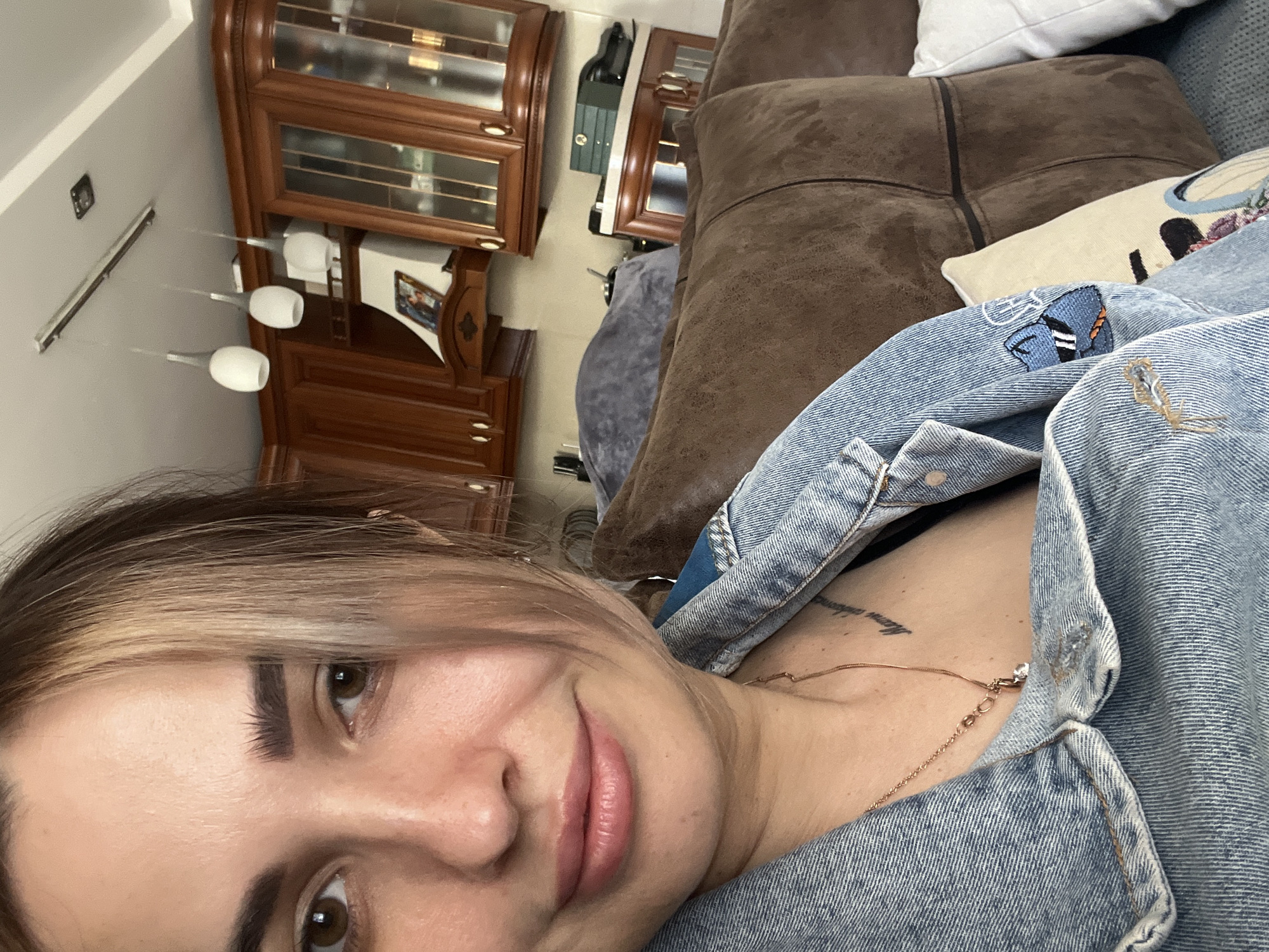 A selfie taken by a woman with light brown hair and fair skin. She is wearing a denim jacket and a gold necklace. The background includes a brown leather couch with pillows, a wooden cabinet, and a modern chandelier. The location appears to be indoors, likely a living room.
