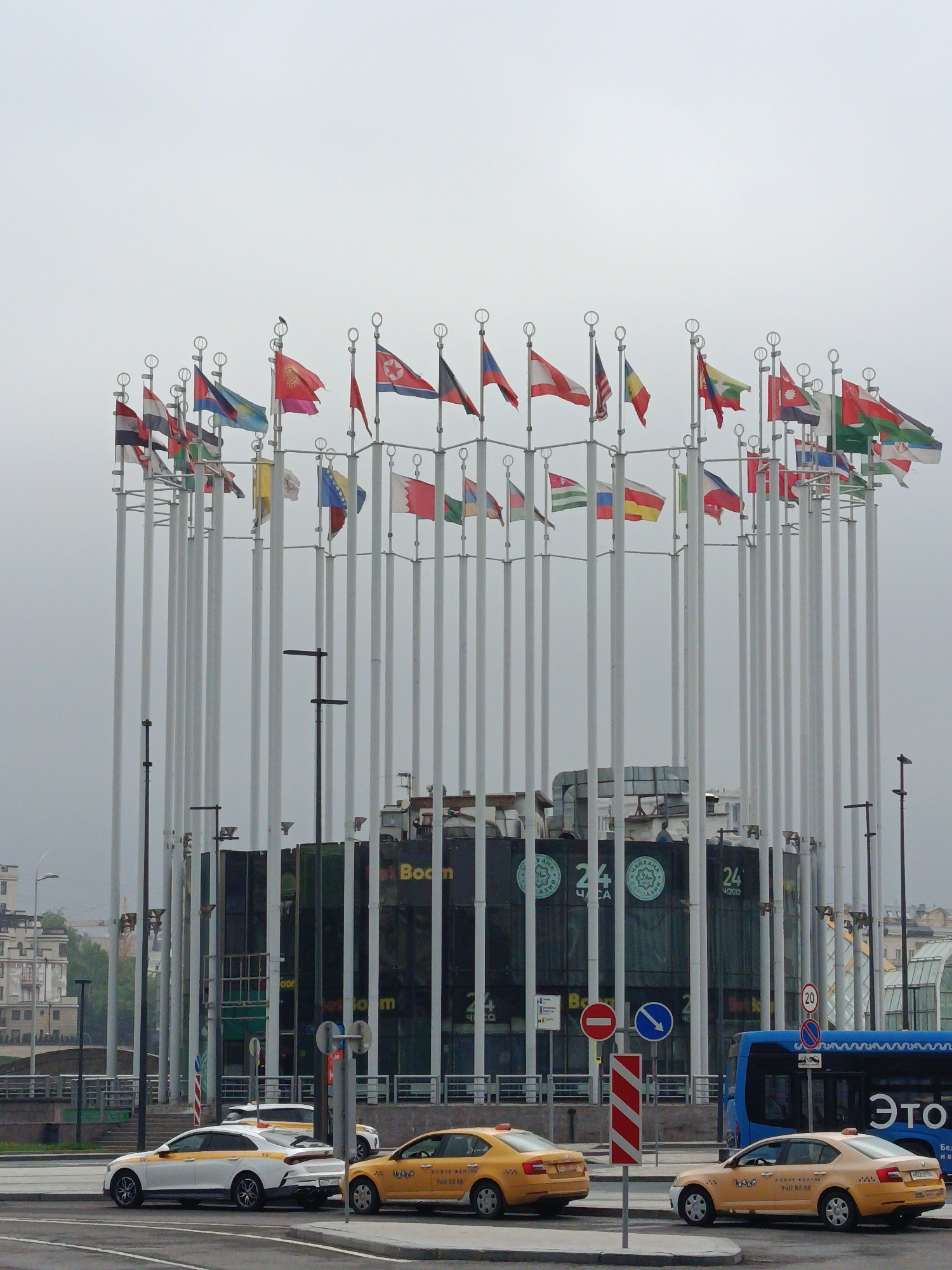 The image captures a street scene in Moscow, Russia, under an overcast sky. In the foreground, three taxis are visible: one white with a yellow stripe and two yellow. A blue bus is also present. A traffic island holds a series of road signs. In the background, a circular arrangement of flagpoles stands, each bearing a different national flag. Behind the flagpoles is a building with "Boom" clearly visible in large letters. The building also features several "24" signs. Streetlights and other buildings can be seen in the far distance.