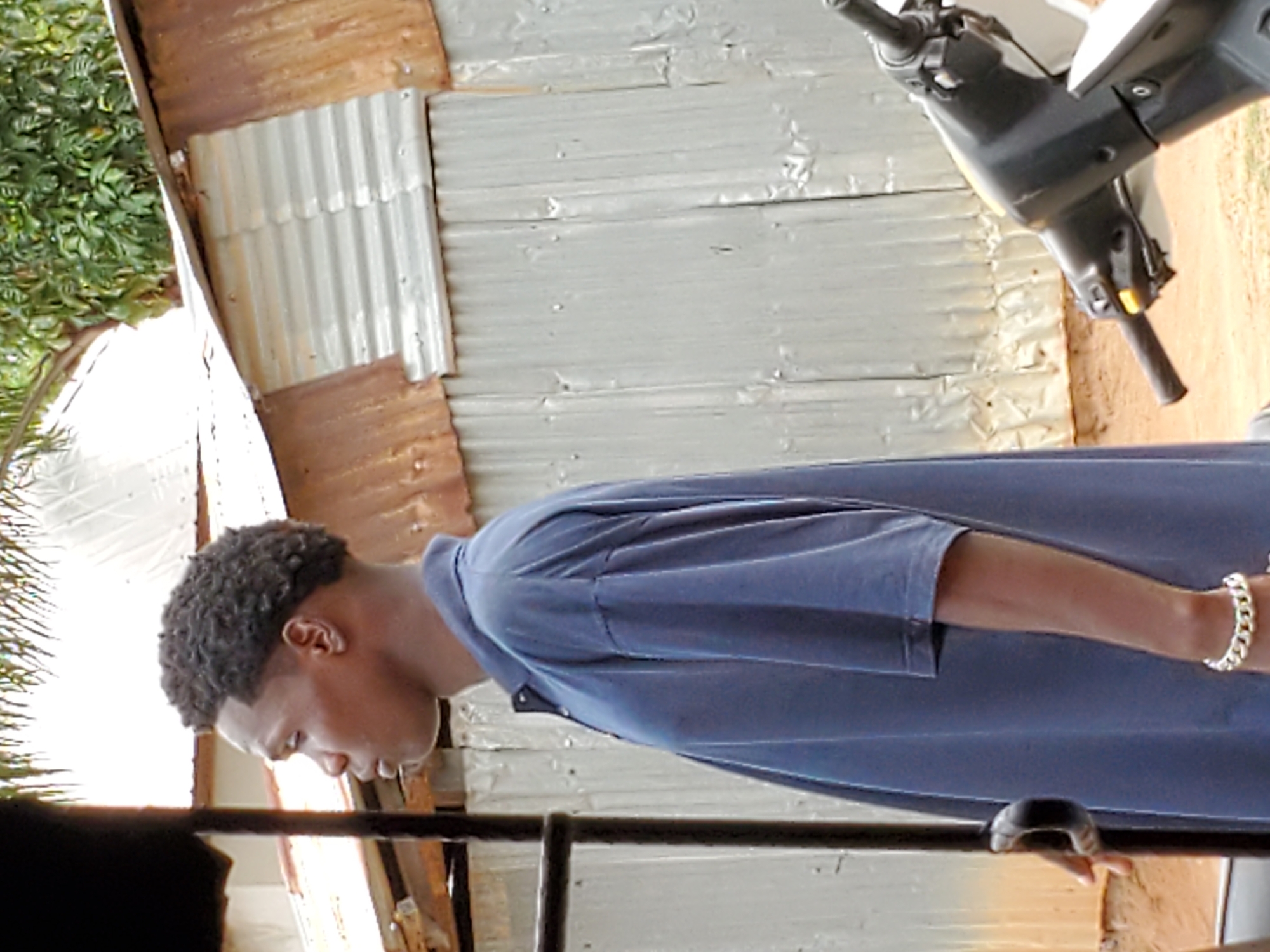 The image shows a man standing near what appears to be a residential structure in Mararaba, Nigeria, based on the corrugated metal siding commonly used in building construction in the region. The man is wearing a blue short-sleeved shirt and a gold bracelet on his left wrist. He has dark hair and appears to be looking down. To the right of the man, partially visible, is a scooter or small motorcycle. The environment consists of the side of the building and possibly foliage in the background.