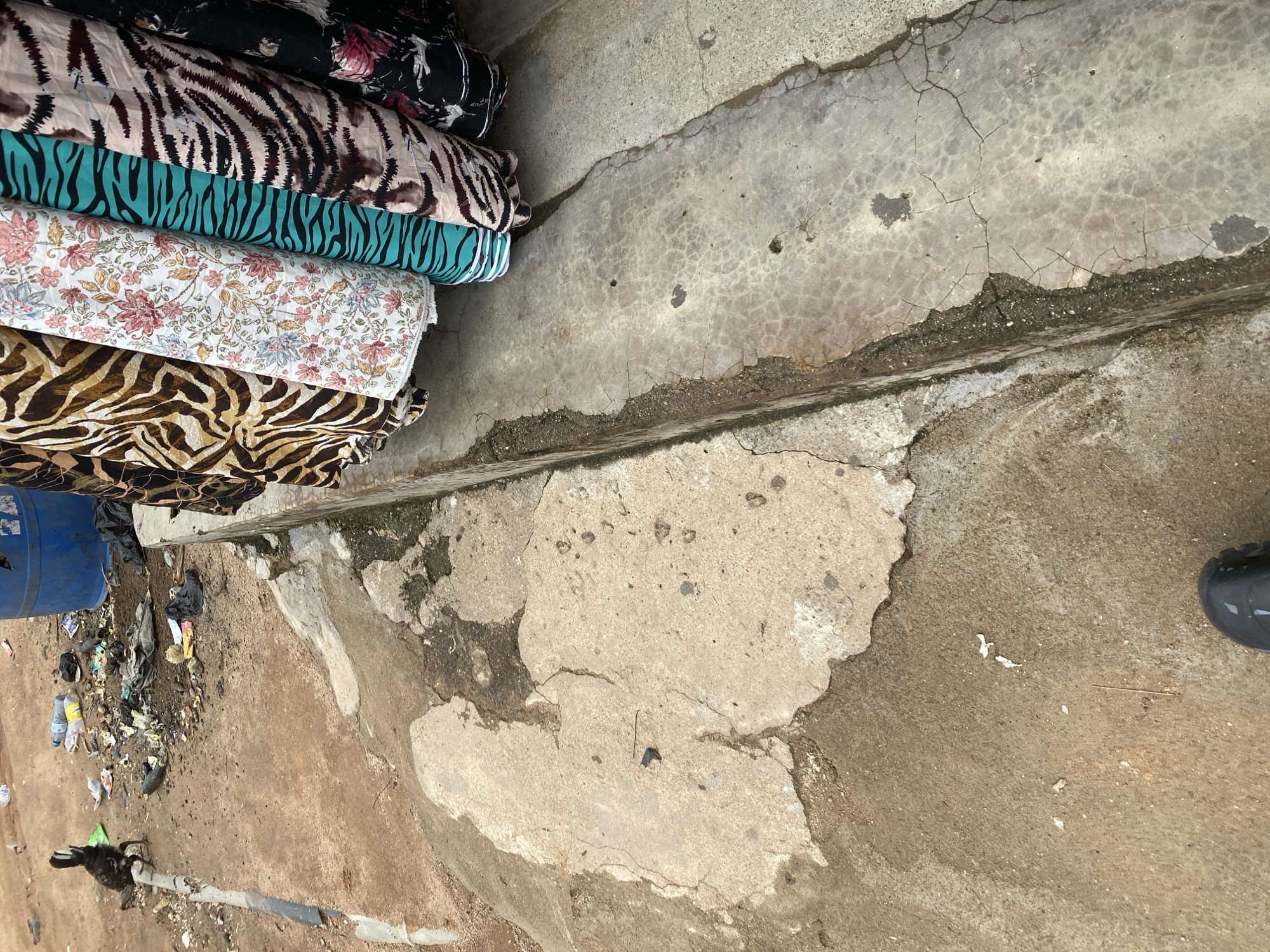 The image shows a stack of fabric rolls, primarily with animal print and floral patterns, placed on the ground next to a concrete surface. There is a blue barrel and scattered trash, including plastic bottles and bags, to the left of the fabric rolls. The ground is a mix of dirt and cracked concrete. A portion of a dark-colored shoe is visible in the lower right corner. The scene appears to be outdoors, possibly a market or roadside stall in Jalingo, Nigeria. The lighting suggests it is daytime. There is no visible text in the image.