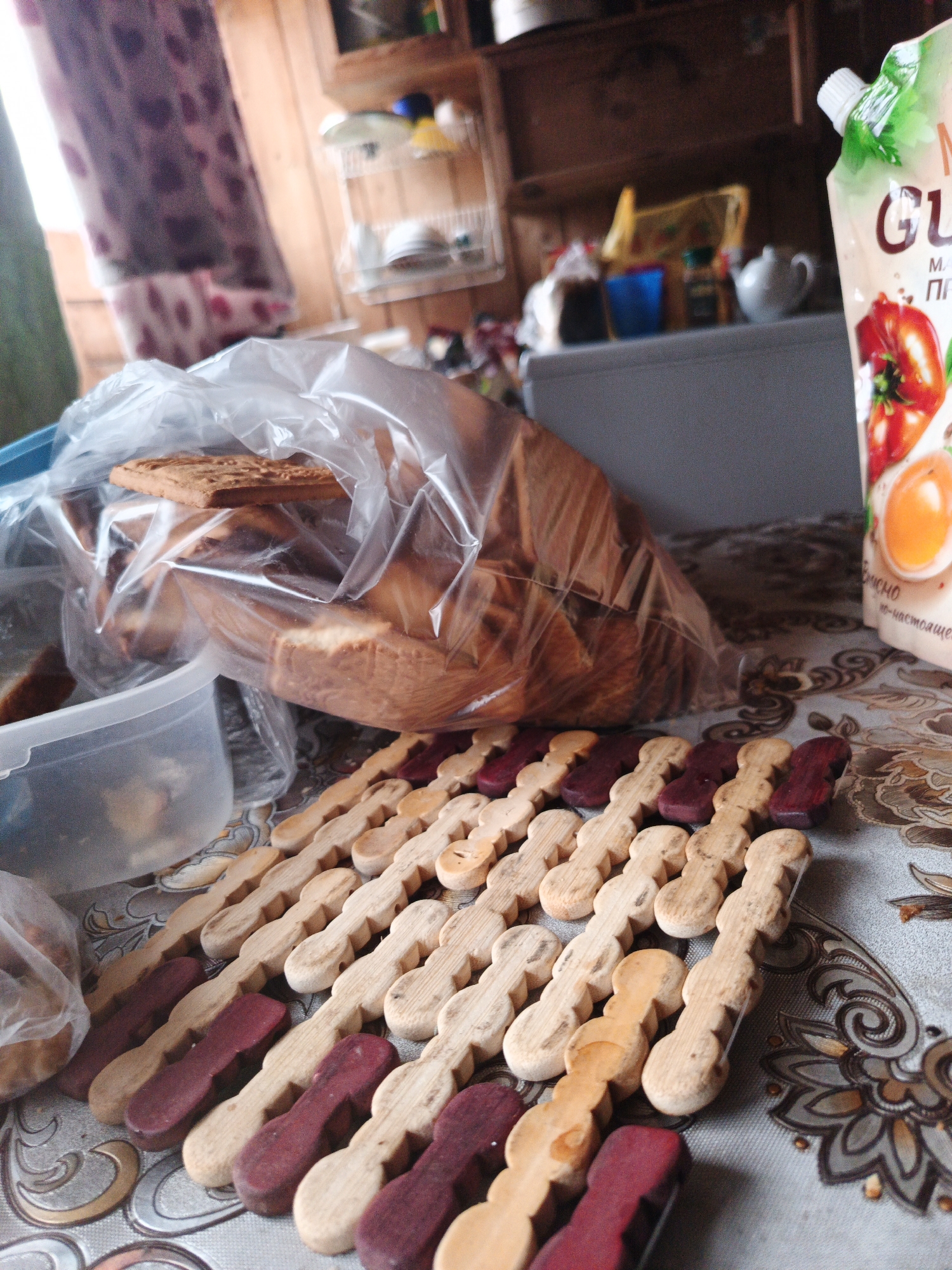 In Islambaevo, Russia, a low-angle, medium shot captures a tabletop setting. A partially opened plastic bag filled with baked goods, possibly cookies or biscuits, rests on a patterned tablecloth. To the right of the bag, a carton of liquid, likely juice, with an image of tomatoes and an egg, is visible. In the foreground, a line of wooden objects, some light brown and some dark red, are arranged in parallel rows. These objects have a distinctive shape, resembling small, elongated figures with rounded ends. In the background, shelves are visible, holding various containers and what appears to be a kettle. The overall impression is one of domestic life.