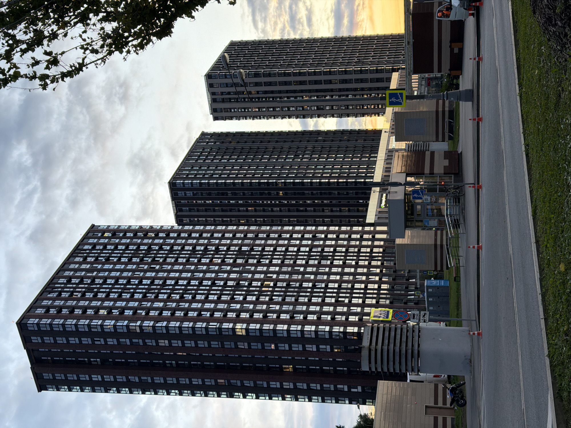 In Khimki, Russia, three towering residential buildings dominate the scene, their dark facades adorned with numerous windows reflecting the overcast sky and the warm hues of a setting sun. In the foreground, a road with double white lines separates the viewer from a grassy area. A pedestrian crossing is marked by a yellow sign with a walking figure. Orange traffic cones are strategically placed on the pavement. In the background, a motorcycle is parked near a large cylindrical structure. Various portable toilets and construction materials suggest ongoing work or maintenance. A white van with an open door and an orange-clad figure is also visible. The overall impression is that of a modern urban landscape during the twilight hours.