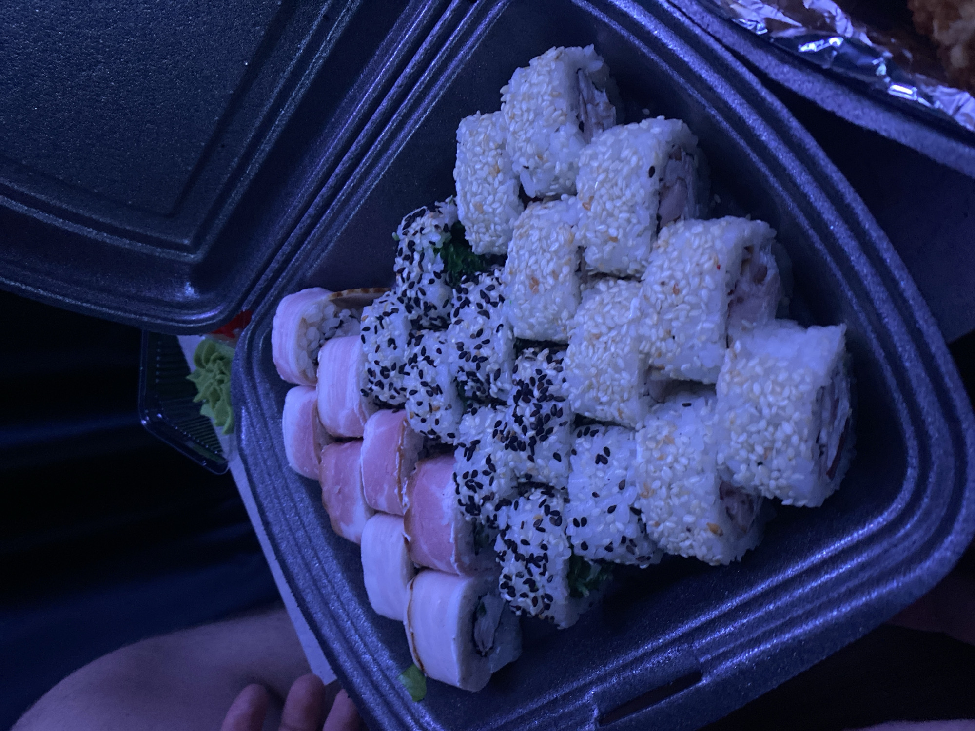 A takeout container filled with sushi rolls sits indoors. The rolls include white sesame-covered, black sesame-covered, and pink-wrapped varieties. Wasabi is visible on the side. The location is unknown. There is no evidence that this is Izhevsk, Russia.
