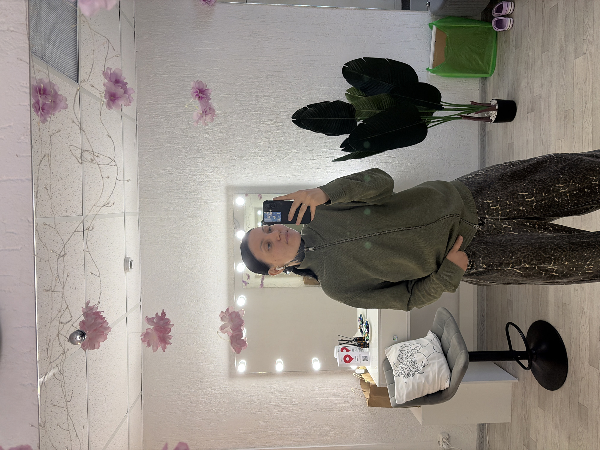 A woman is taking a selfie in a room, likely a studio, in Chaikovsky, Russia. The room has a large mirror with lights around the perimeter, creating a vanity area. The wall behind the mirror is white with floral decorations. The woman is wearing a green zip-up jacket and patterned pants. To the right of the woman in the reflection is a potted plant, a green shopping bag, and a pair of sandals on the floor. In front of the mirror is a white desk with a grey chair and a cushion on it. On the desk are various items, including bottles, packages, and a decorative object. The floor is light-colored wood.