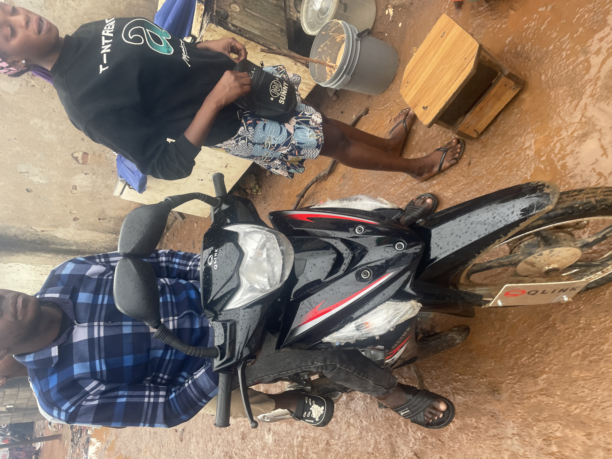 The image depicts a street scene in Kurunduma, Nigeria, on a wet day. Two individuals are present. A person wearing a blue and black plaid shirt and dark pants is seated on a black motorcycle. The motorcycle has red and white accents and appears to be parked on a muddy street. To the right of the motorcycle, a person wearing a black sweatshirt with white lettering and a patterned skirt is standing. This person is holding a black pouch with "SUNNY" printed on it. Nearby, there is a grey bucket with a wooden stick inside, and a small wooden stool. The ground is covered in mud and puddles, suggesting recent rainfall. The background shows a weathered wall of a building.