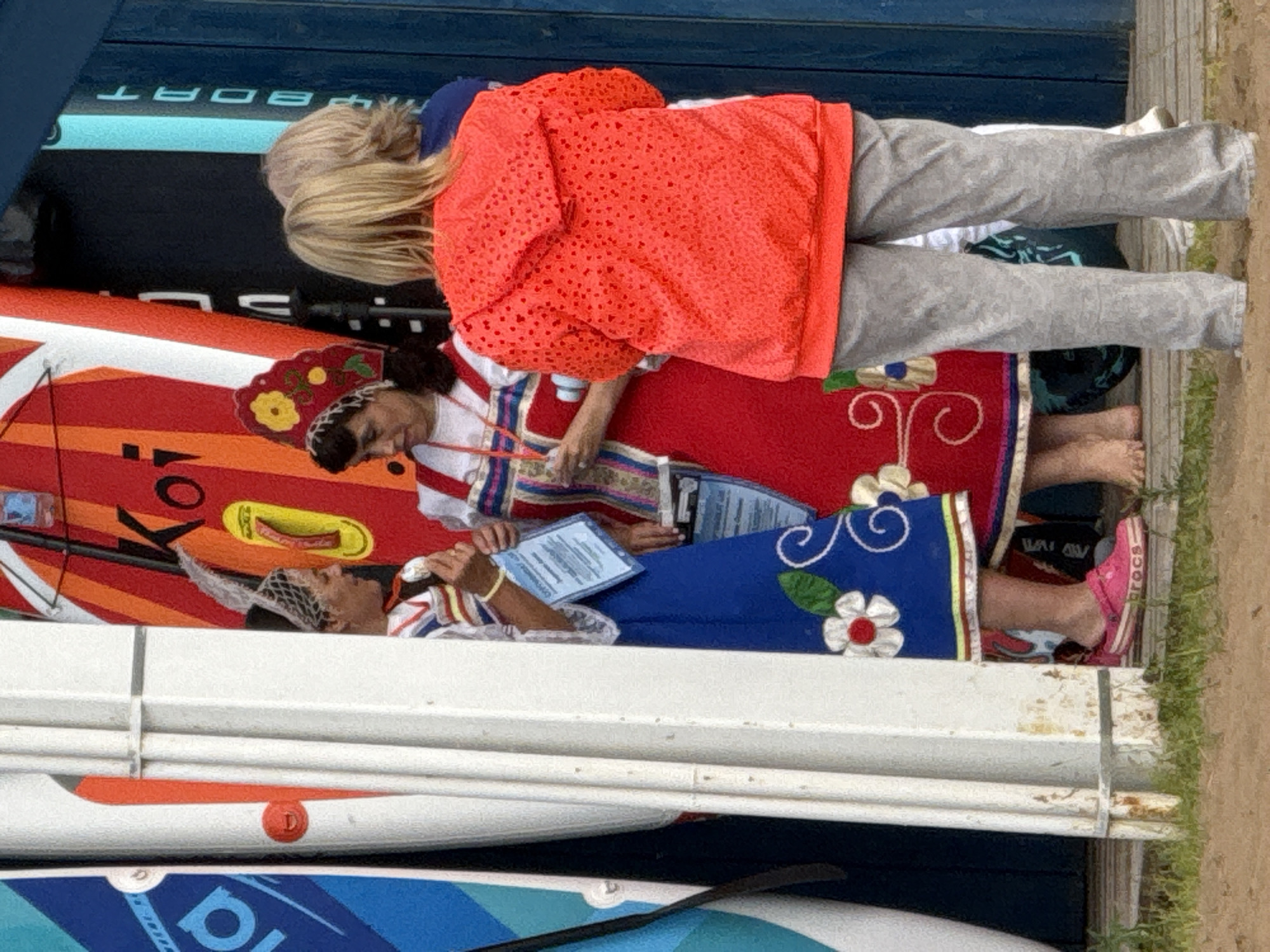 The image depicts an outdoor scene with several individuals standing near a colorful display of what appear to be paddleboards or surfboards. Two women in traditional Russian folk attire, characterized by embroidered dresses and headdresses, are positioned to the left. One woman, wearing a blue dress with white floral embroidery, is holding a blue certificate or document. The other woman, in a red dress with similar folk embellishments, is also holding a similar document. Their posture and the documents suggest they might be participating in an event or ceremony.
Behind them, a person wearing a bright orange hooded jacket with a patterned texture is visible, facing away from the camera. Another individual, whose back is to the viewer, is wearing a blue shirt. The backdrop consists of vertical elements that could be part of a structure or the boards themselves, painted in vibrant colors like red, orange, and blue, with some text visible on the blue board to the far right that reads "BOAT".
The ground in the foreground is a sandy or dirt surface with some green grass. The overall atmosphere suggests a public gathering or festival, possibly related to water sports given the presence of the boards. The scene is illuminated by natural light, indicating it is daytime. The image appears to have been taken in the city of Chaikovsky, Russia, possibly at a waterfront location or a venue hosting an event.