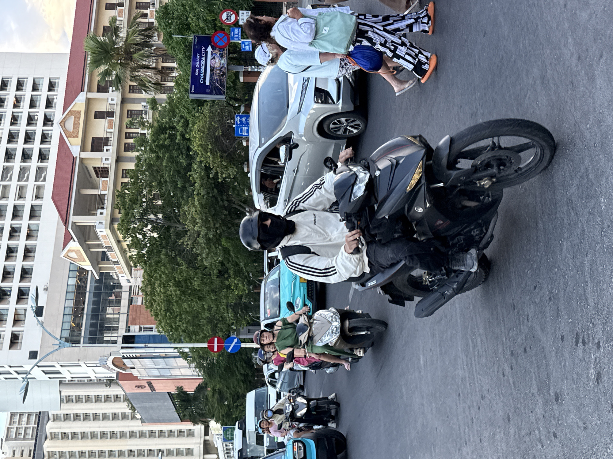 In Nha Trang Ward, Vietnam, a street scene unfolds with a moderate traffic flow. A person wearing a black mask and a white jacket with black stripes rides a black motorcycle in the foreground, heading towards the viewer. Behind them, a silver minivan and a turquoise car are visible, alongside other motorcycles with multiple passengers. Pedestrians, including two women, one in a white top and patterned pants and the other in a white shirt and striped pants, walk on the right side of the street. The environment includes tall buildings, lush green trees, street signs, and traffic signals, suggesting an urban setting. The lighting indicates it is daytime, possibly late afternoon.