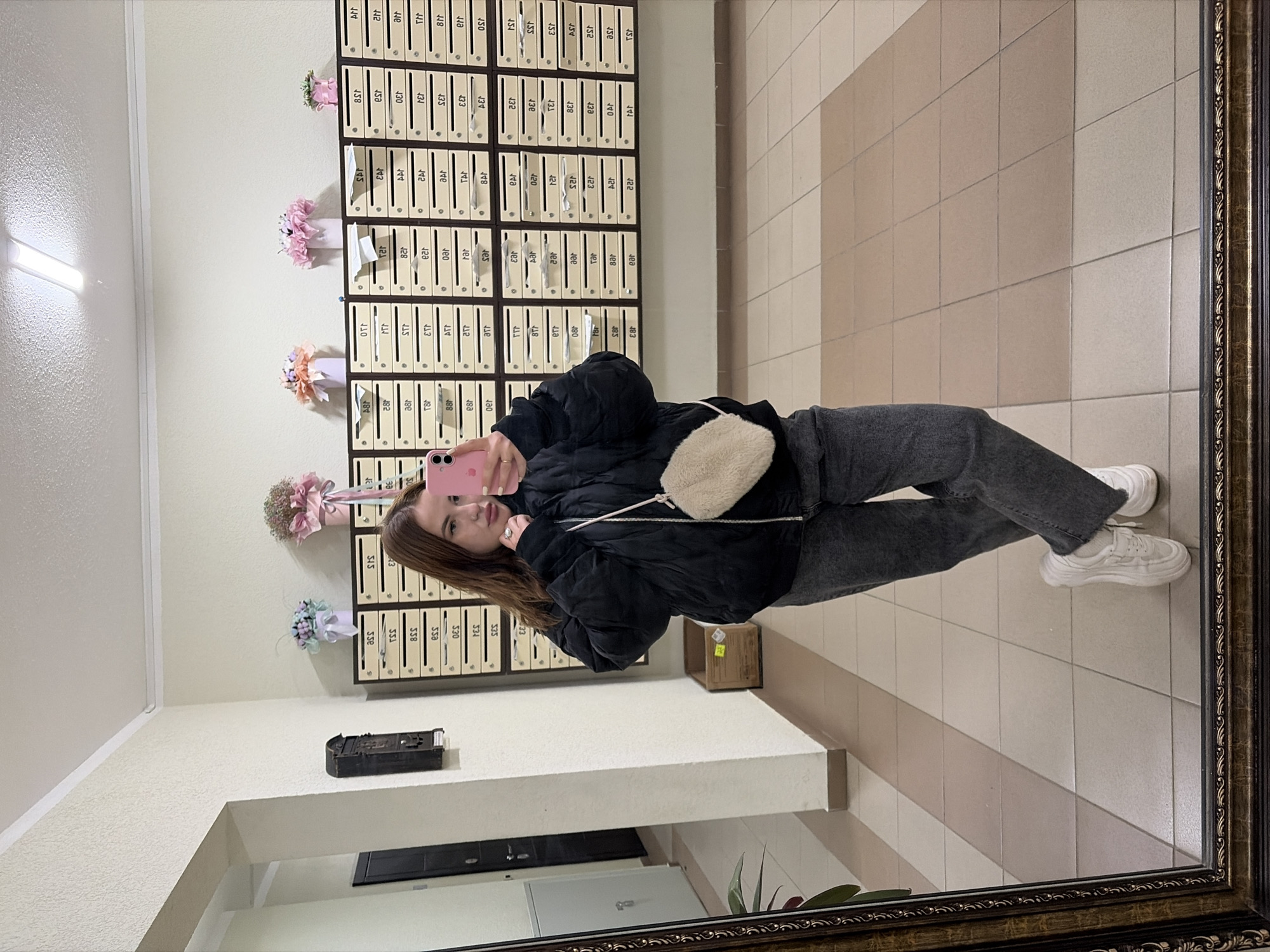A young woman is depicted in a mirror selfie taken indoors, likely in an apartment building in Krasnodar, Russia. She stands upright, holding a pink smartphone with both hands to capture her reflection. She has long brown hair and is dressed in a black puffer jacket, dark grey jeans, and white sneakers, with a small, light-colored fluffy crossbody bag hanging from her shoulder. Her expression is calm and posed, with a slight smile.
The setting is an indoor hallway or common area. Behind the woman, a wall is covered with numerous light-colored mailboxes arranged in an extensive grid, each featuring dark, three-digit numbering. Small decorative floral arrangements in pink, light blue, and purple vases are placed on top of some of the mailboxes, adding a touch of color to the otherwise functional space. The floor is tiled with alternating light and darker beige/brown square tiles, creating a patterned surface. A long fluorescent light fixture on the light-colored, textured ceiling illuminates the area.
Other reflected elements include a dark, ornate object (possibly an old mailbox or decorative shelf) on a ledge to the left, a small cardboard box on the floor to the right, and a white appliance (potentially a refrigerator or locker) partially visible below the ledge on the left, accompanied by some greenery. The mirror itself is enclosed by an ornate dark brown frame with golden accents.
Visible text, primarily from the mailboxes, includes numerous three-digit numbers such as 158, 140, 133, 128, 123, 110, 101, 240, 233, 220, 211, 201, 345, 334, 320, 310, 301, 448, 430, 420, 410, 401, 558, 530, 520, 510, 501, and many more. These numbers often appear reversed due to the mirror reflection. Some reversed letters like "SAT" and "AST" are also present.