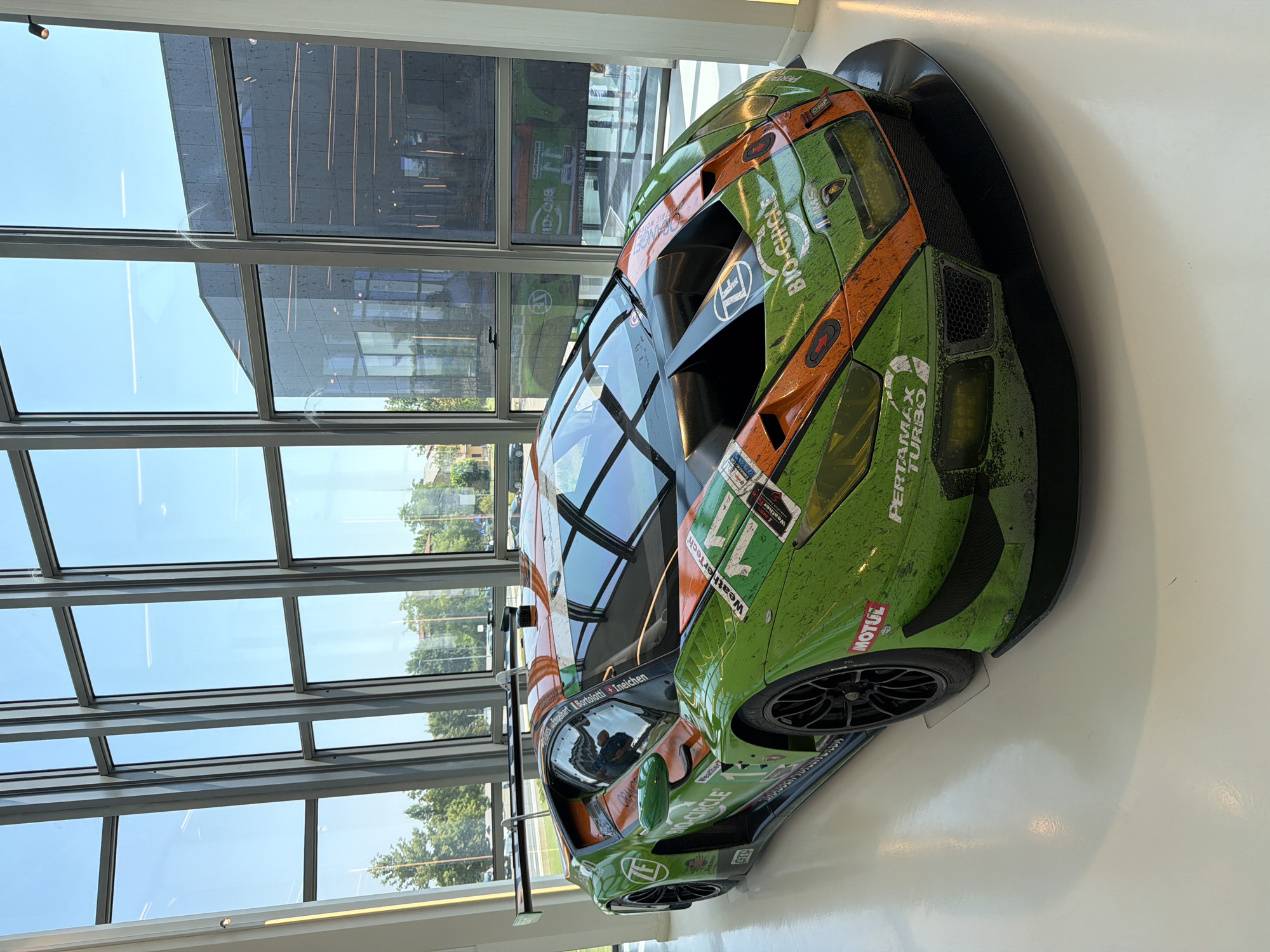 A green and orange racing Lamborghini, covered in dirt and debris, is parked indoors on a polished white floor. The car features several sponsor logos, including "ZF", "BIO-CIRCLE", and "PERTAMAX TURBO". Behind the car is a large wall of windows that looks out onto a tree-lined street and buildings. The reflection in the windows shows the interior of the building, which appears to be a showroom or museum, with more windows and architectural elements. The lighting in the scene is bright, suggesting daylight. The image was taken in the city of Maggi, Italy.