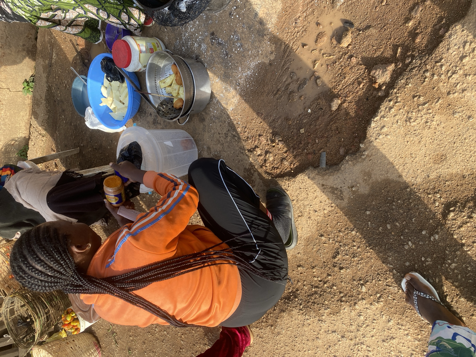 The media file shows an overhead view of a person sitting on the ground in an outdoor setting, likely in Jos, Nigeria. The person is wearing an orange hooded sweatshirt with blue and white stripes on the sleeves and black athletic pants with white stripes. They have long, braided black hair that trails down their back. The person is holding a small yellow jar in their right hand.
In the foreground, to the left of the person, are several woven baskets. One basket contains small, colorful fruits. Another appears empty. Behind the person, there are various kitchen items arranged on the ground. These include a large blue bowl filled with cut white produce, likely tubers, and a smaller blue bowl containing black items, possibly plastic bags. A metal colander holds fried food, possibly plantains or yams, and a large white jar with a red lid is also visible. A clear plastic container is also present.
The ground is a mix of dirt, gravel, and a muddy patch, suggesting it might be a street or an unpaved area. There are some scattered white particles on the ground, which could be flour or dust. Sunlight casts shadows across the scene, indicating it is daytime. The overall impression is of a casual, possibly commercial setting, where food preparation or sale might be taking place. There is no visible text in the image.