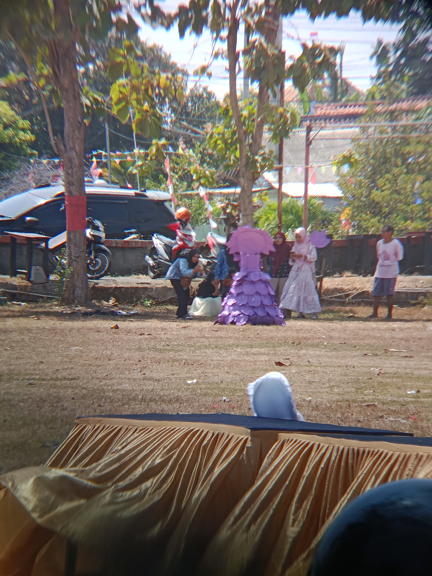 The image captures a scene outdoors, possibly in Desa Kebon Manis, Kecamatan Cilacap Utara, Indonesia, under a bright sky. In the foreground, a table draped with a gold, pleated cloth is prominent, with a dark object, possibly a person's head or a piece of equipment, partially visible in the bottom right corner. In the mid-ground, dry, grassy ground is visible. A group of people are gathered. A person in a white hijab is seated on the ground to the left, partially obscured by the table. Further back, a person in a purple, layered costume resembling a flower or a bell stands center, with two other people in hijab standing to their right. To the far right, a person in a pink t-shirt and blue shorts stands. In the background, a dark-colored SUV is parked, with motorcycles nearby. Trees with green foliage are present throughout the scene, and small, colorful flags are strung across the area, suggesting an event or celebration. The overall impression is of a daytime outdoor gathering.