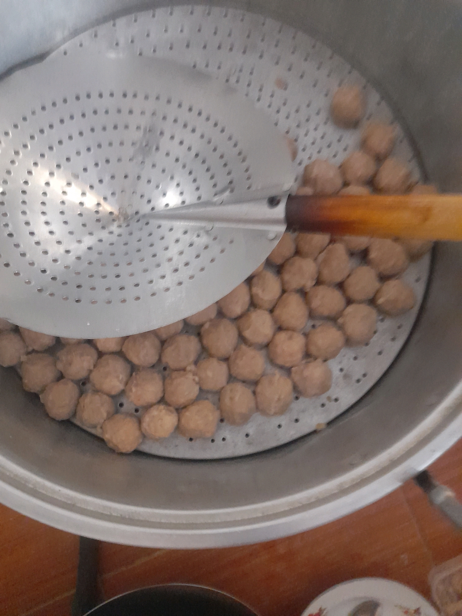 The image shows a close-up, high-angle view of a metal steamer pot filled with numerous round, light brown meatballs. A silver, perforated steamer basket is positioned inside the pot, holding the meatballs. A wooden-handled scooping utensil with a silver, perforated head rests on the edge of the steamer basket, partially covering some of the meatballs. The pot appears to be on a table with an orange, textured surface, possibly wood or tile. The background is blurred, but suggests an indoor environment with some light sources. The image does not provide any specific information or context that would allow for the determination of a location in Pucangro, Indonesia.