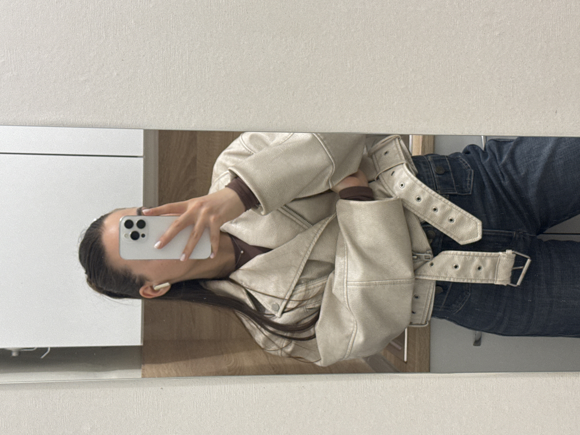 A woman is reflected in a mirror while taking a selfie with a white smartphone. She is wearing a light beige, faux leather jacket and dark denim jeans. She has brown hair and is wearing a white earbud. The background reflection includes elements of what appears to be a simple interior, possibly a home or apartment, with white walls and wood accents. The location in Sofia, Bulgaria, cannot be determined from the image.
