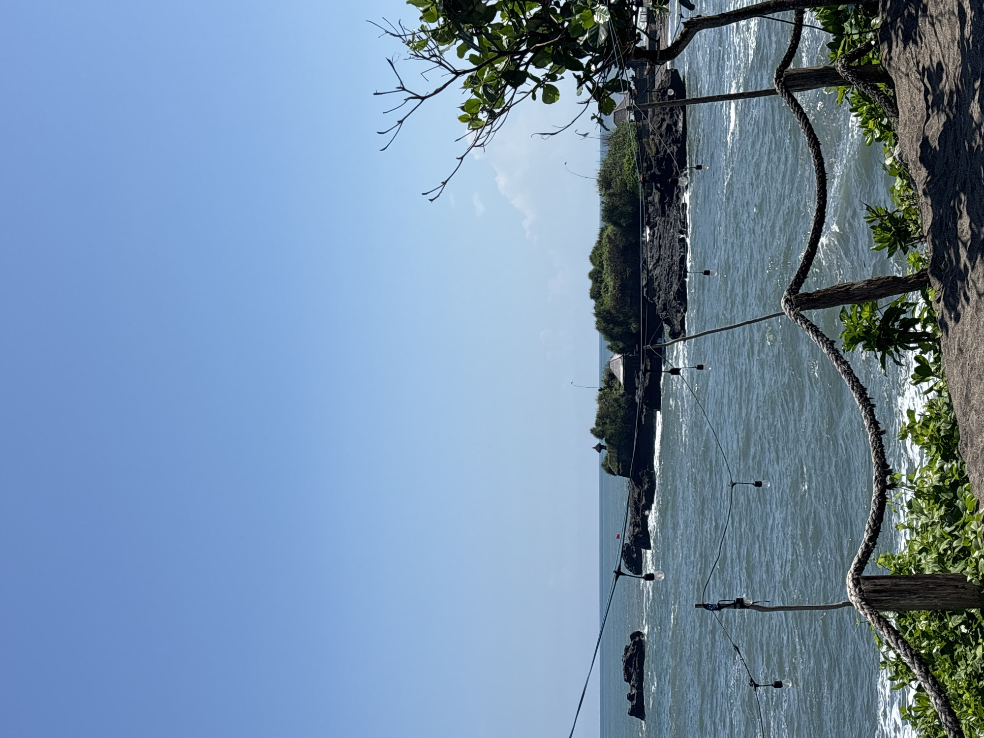 A seaside view is presented, likely in Mengwi, Indonesia, as suggested by the location. The left side of the image is dominated by a clear, blue sky. A rocky coastline is visible in the center, covered with dense greenery. The sea stretches towards the horizon. A decorative rope fence with light bulbs runs along the right side of the frame, with leaves and a tree branch visible at the top.