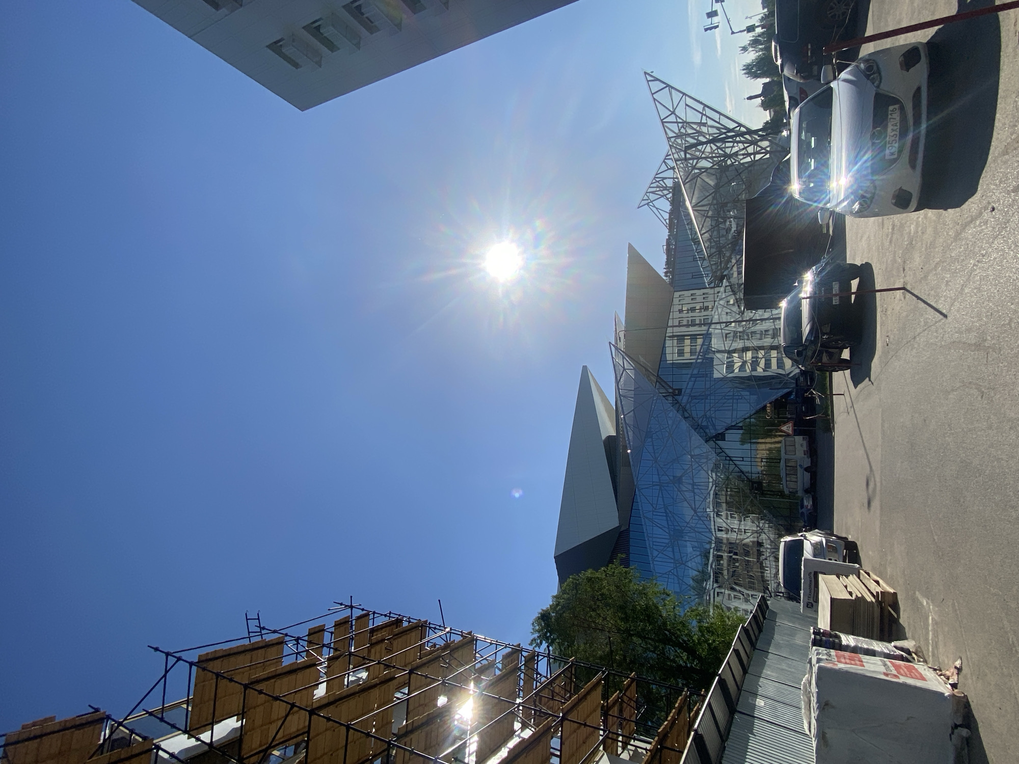 The image shows a view in Kazan, Russia, looking upwards towards a modern glass building with a sharp, angular design. The sun is high in the clear blue sky, causing a bright glare. Partially obscuring the view is scaffolding, likely part of an adjacent construction or renovation project. Several cars are parked on the street, and construction materials, including wrapped stacks, are visible along the curb. The buildings are of modern construction.