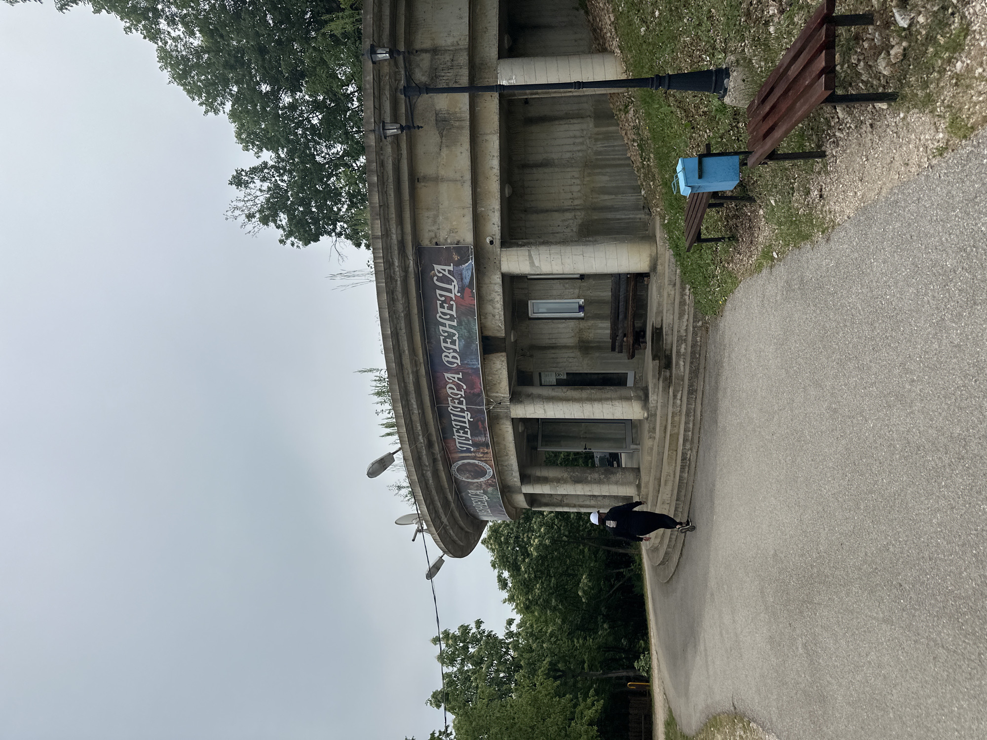 The image shows a partially obscured view of "Пещера Венеца" (Peshtera Venetsa), a cave in the Gara Oreshets region of Bulgaria. The cave entrance is a semicircular, concrete structure with columns. A banner hangs on the structure, displaying the cave's name in Bulgarian. The sky is overcast. A person, dressed in dark clothing, is walking away from the entrance on a paved path. A bench and waste bin are located to the side of the path. Trees surround the area.
