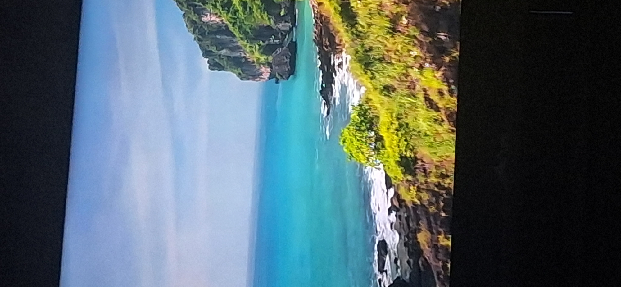 The image depicts a scenic coastal landscape with a clear blue sky and turquoise ocean. A rocky cliff covered in lush green vegetation rises from the water on the right side of the frame. The shoreline in the foreground is characterized by dark, jagged rocks and white-capped waves. The overall scene evokes a sense of natural beauty and tranquility.
The provided image does not contain any elements or features that can be used to identify a location within the city of Bila Tserkva, Ukraine, or any other specific geographical area.