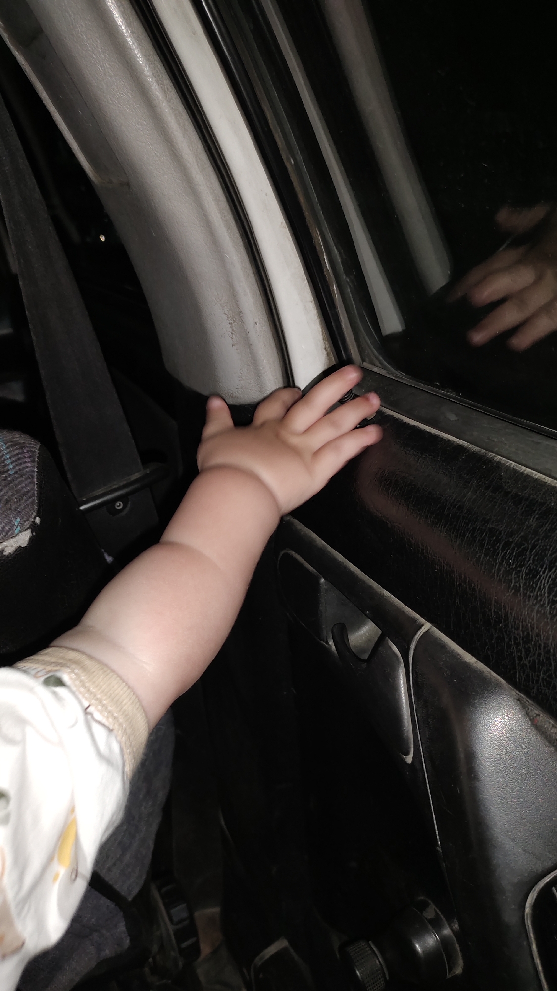A close-up shot focuses on a baby's arm and hand reaching out toward the window of a car. The baby's skin is soft and slightly flushed, and their fingers are spread open as they touch the edge of the window frame. The arm is clad in a light-colored long-sleeved shirt with a subtle pattern of yellow and white shapes.
The car's interior is visible, with a black leather door panel featuring a silver-toned door handle. A black seatbelt is also prominently featured on the left side of the frame, along with a portion of the car seat. The window appears to be slightly tinted, and a reflection of another hand, presumably an adult's, can be seen in the glass.
The lighting suggests it may be nighttime or a dimly lit environment, casting shadows on the car's interior. The overall impression is one of a quiet, everyday moment, possibly during a car ride, where the baby is exploring their surroundings. No visible text is present in the image.