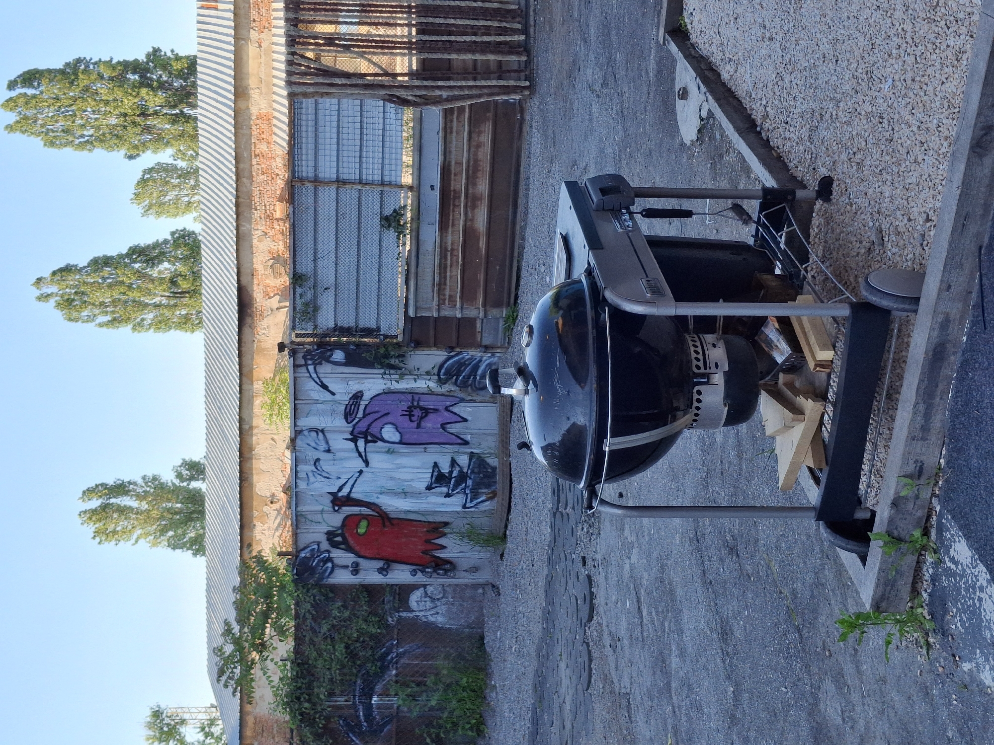 The image shows a Weber grill on a paved surface in an outdoor urban environment. Behind the grill is a dilapidated building facade with graffiti, including two cartoonish characters painted on a weathered wooden door. The building has a corrugated metal roof and a brick structure with barred windows. Vegetation grows along the base of the building. Trees are visible in the background against a clear blue sky. A gravel area is partially visible to the right of the grill, bordered by wooden planks.
