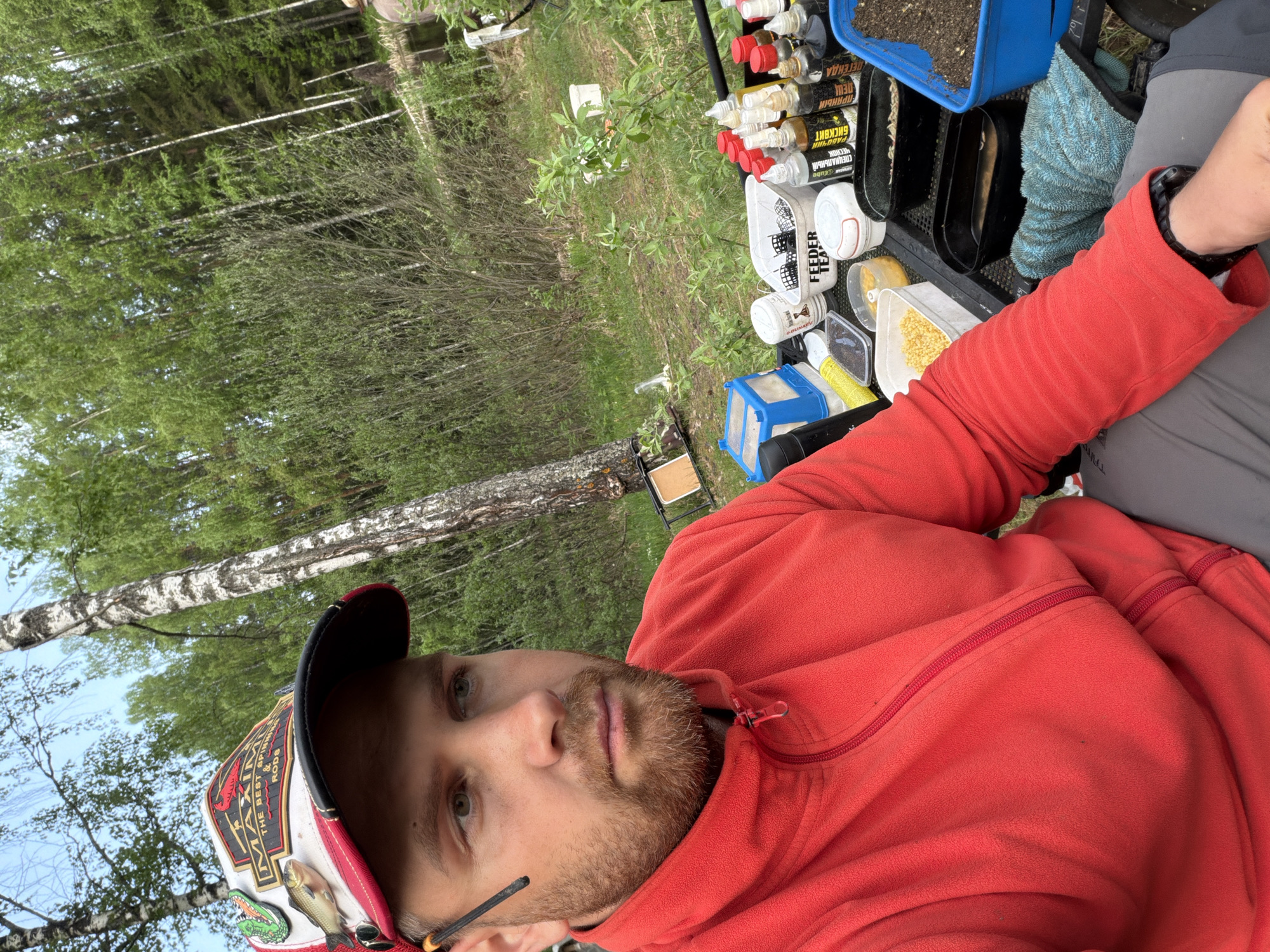 A man with a beard, wearing a red fleece jacket and a cap, is sitting outdoors. The cap has a logo with the text "THE BEST SPINN RODS &". Patches of a fish and an alligator are attached to the cap. To the right of the man, various fishing supplies are arranged in trays and containers, including bottles, boxes, and a container of corn. The labels on the bottles are partially visible, with some words in Russian. The background shows a forest landscape with birch trees. The OCR text suggests the products are related to fishing, potentially including brands and flavors. The location is Chaykovsky, Russia.