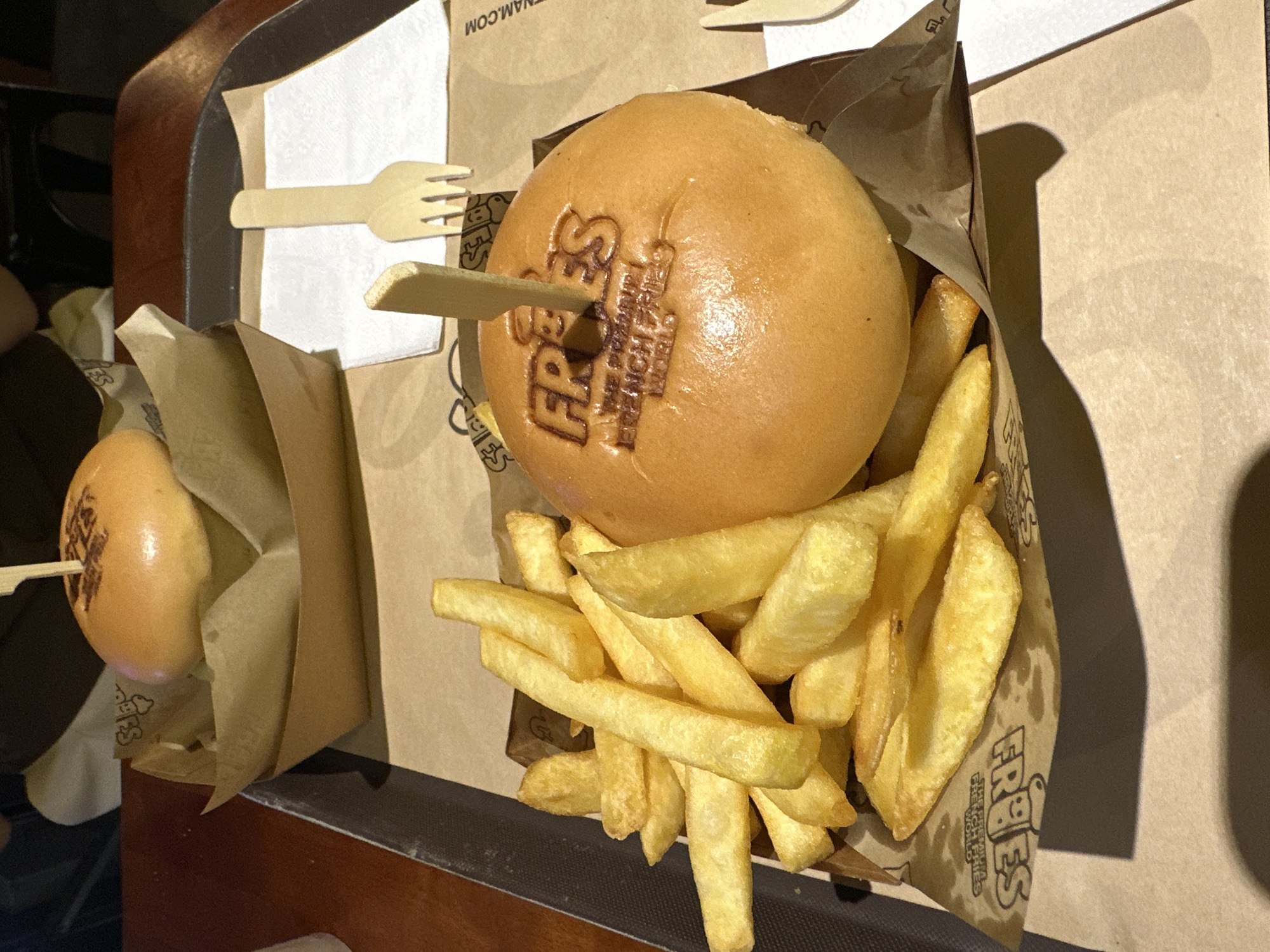 A tray presents two servings of "FRIES" branded food, possibly in Thủ Đức, Vietnam (city inferred from other examples). Each serving consists of a small round bun with "FRIES" imprinted on top, secured with a wooden skewer, and a portion of thick-cut French fries contained in a paper bag. A disposable wooden fork rests on a white napkin within the tray. The food is illuminated by warm lighting.