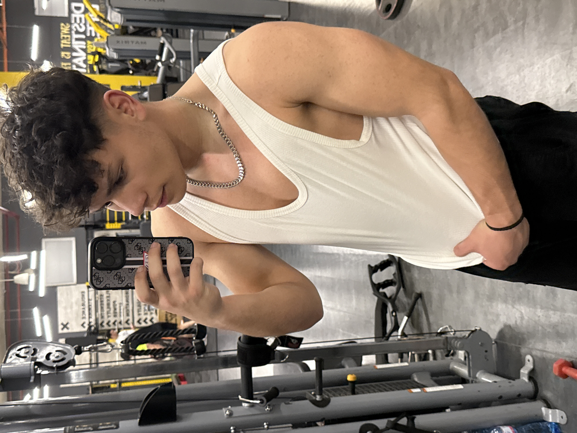 The image depicts a young man taking a selfie in what appears to be a gym. He is wearing a white tank top and black pants, and a silver chain around his neck. He is holding a smartphone with a patterned case. Exercise equipment is visible in the background. Given the presence of Cyrillic text and what appears to be a Bulgarian weight plate, the location is likely in Sofia, Bulgaria.