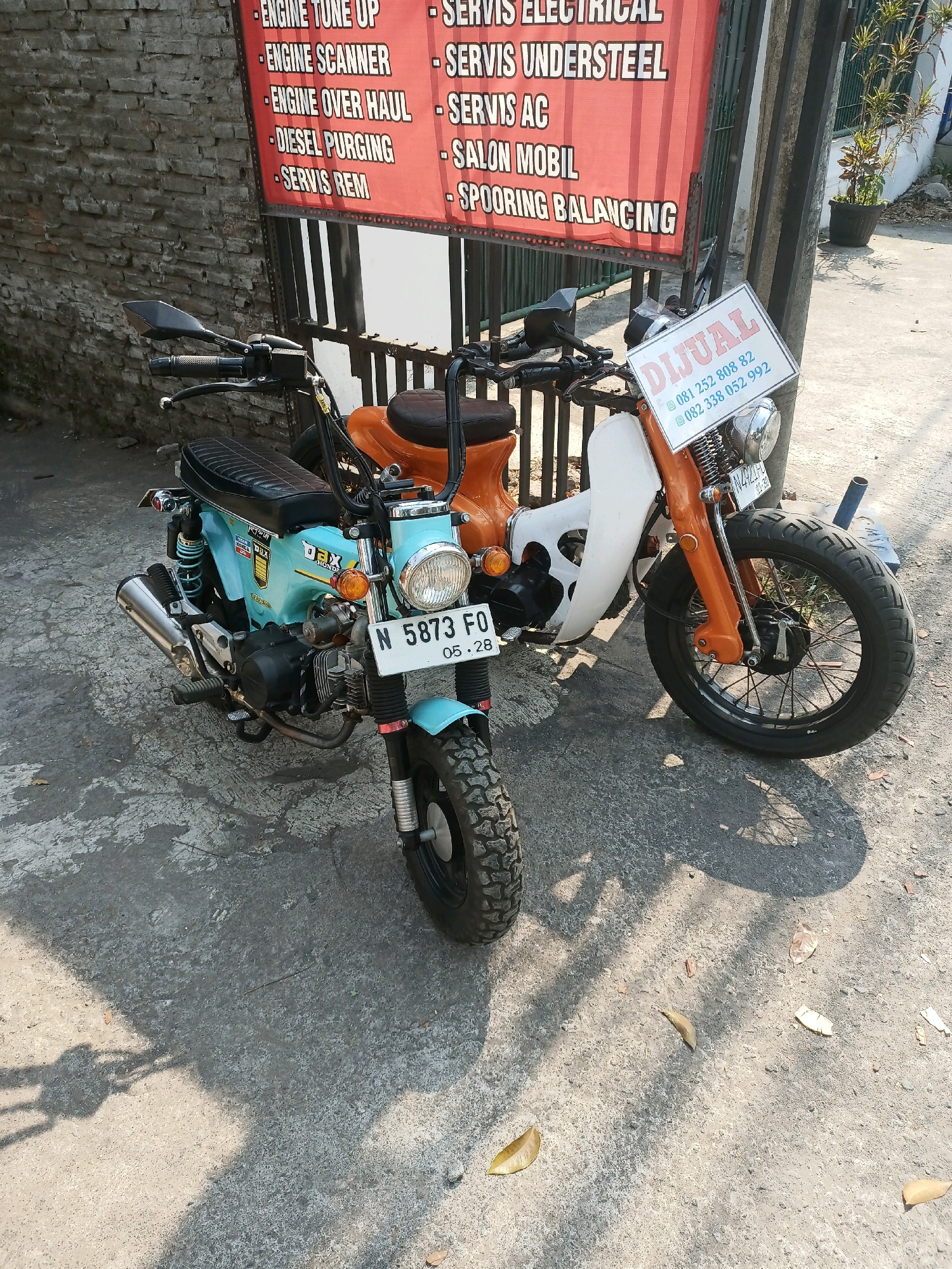 Two motorcycles are parked on a paved surface, casting shadows from the sunlight. In the background, a brick wall stands to the left, and a metal fence separates the parking area from a building with a red banner displaying white text. The banner lists various services, including "ENGINE TUNE UP," "ENGINE SCANNER," "ENGINE OVER HAUL," "DIESEL PURGING," "SERVIS REM," "SERVIS ELECTRICAL," "SERVIS UNDERSTEEL," "SERVIS AC," "SALON MOBIL," and "SPOORING BALANCING." To the right of the fence, a potted plant is visible near a light pole. A white sign with red and blue text is attached to the handlebars of the motorcycle on the right. This sign reads "DIJUAL" (For Sale) and provides two phone numbers. The motorcycle on the left is light blue with black accents and a silver exhaust pipe. The motorcycle on the right is orange and white with black handlebars and a black seat. The license plate of the motorcycle on the left is visible and reads "N 5873 FO 05.28." The license plate of the motorcycle on the right is partially obscured, but visible text includes "N 4923." The scene appears to be an outdoor area in a city, potentially a street or a workshop, in Malang, Indonesia, suggested by the "N" license plate prefix common in East Java, Indonesia.