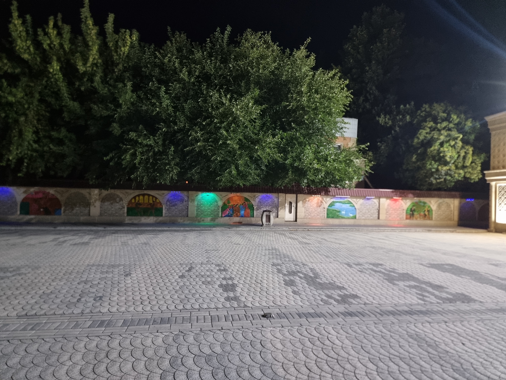 The image depicts an outdoor paved area at night, likely in a city. A patterned stone walkway extends into the foreground. Behind the walkway, a wall adorned with arched niches containing colorful murals is visible. These niches are illuminated by colored lights. Lush green trees are visible behind the wall, with a dark night sky above. In the middle ground, a trash receptacle and a doorway are situated near the wall. The overall scene suggests an outdoor public space, possibly a park or courtyard, decorated with artistic elements.