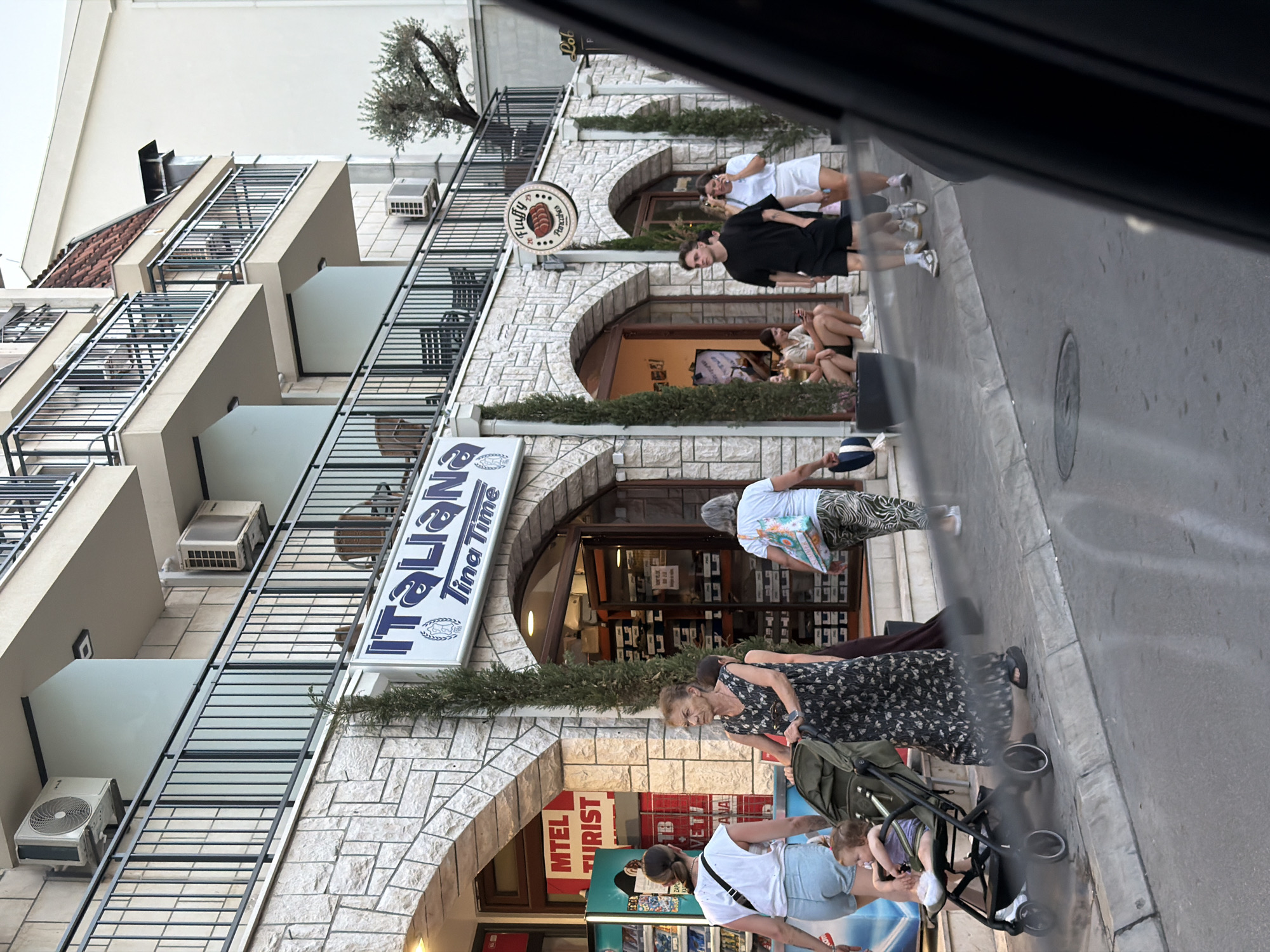 The image depicts a street scene in Budva, Montenegro, featuring a building with multiple levels, balconies, and storefronts. On the ground floor, a business with a sign that reads "Italiana Tina Time" is visible. Adjacent to it, another store with a sign that partially reads "MTEL TURIST" is present. Several people are seen walking along the sidewalk. A woman pushes a stroller with a child in it, while another woman walks beside her. Further down the street, a group of people are seated outside a cafe or shop. The building's facade is made of stone, and there are numerous air conditioning units mounted on the exterior walls. A tree is visible in the background. The street appears to be paved, and there is a sidewalk with a curb. The overall impression is of a commercial area in a European city.