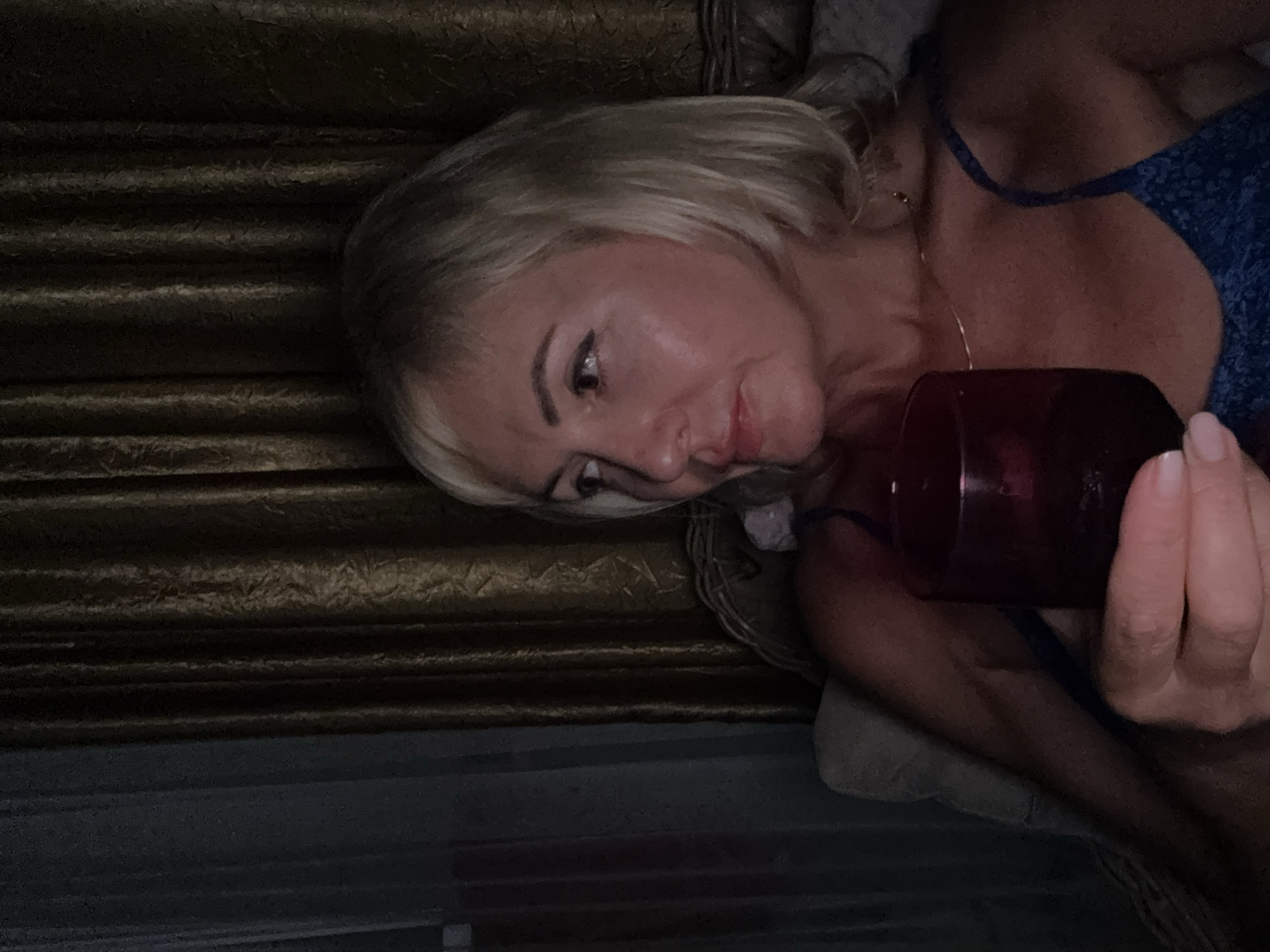 In Alanya, Turkey, an individual with blonde hair and fair skin, wearing a blue garment, is holding a dark red glass filled with liquid. The person is seated, with their head turned to the right. They are indoors, with patterned curtains forming the backdrop. The lighting is dim.
