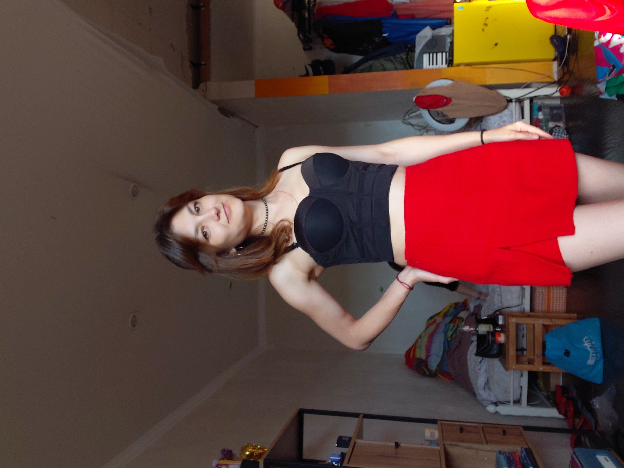 A young woman with brown hair and fair skin stands in a cluttered room, posing for a photo. She is wearing a black corset-style top and a red mini-skirt with a slit. Her left hand is on her hip, and her right arm is bent, with her hand resting on her waist. She is looking directly at the camera with a slight smile. The room appears to be a bedroom, with a made bed visible in the background, covered in colorful blankets. There are shelves with various items on the left side of the room, and clothes are hanging on a rack on the right side. The overall impression is one of a casual, personal space. It is not possible to determine the specific location within Magnitogorsk, Russia, from the image.