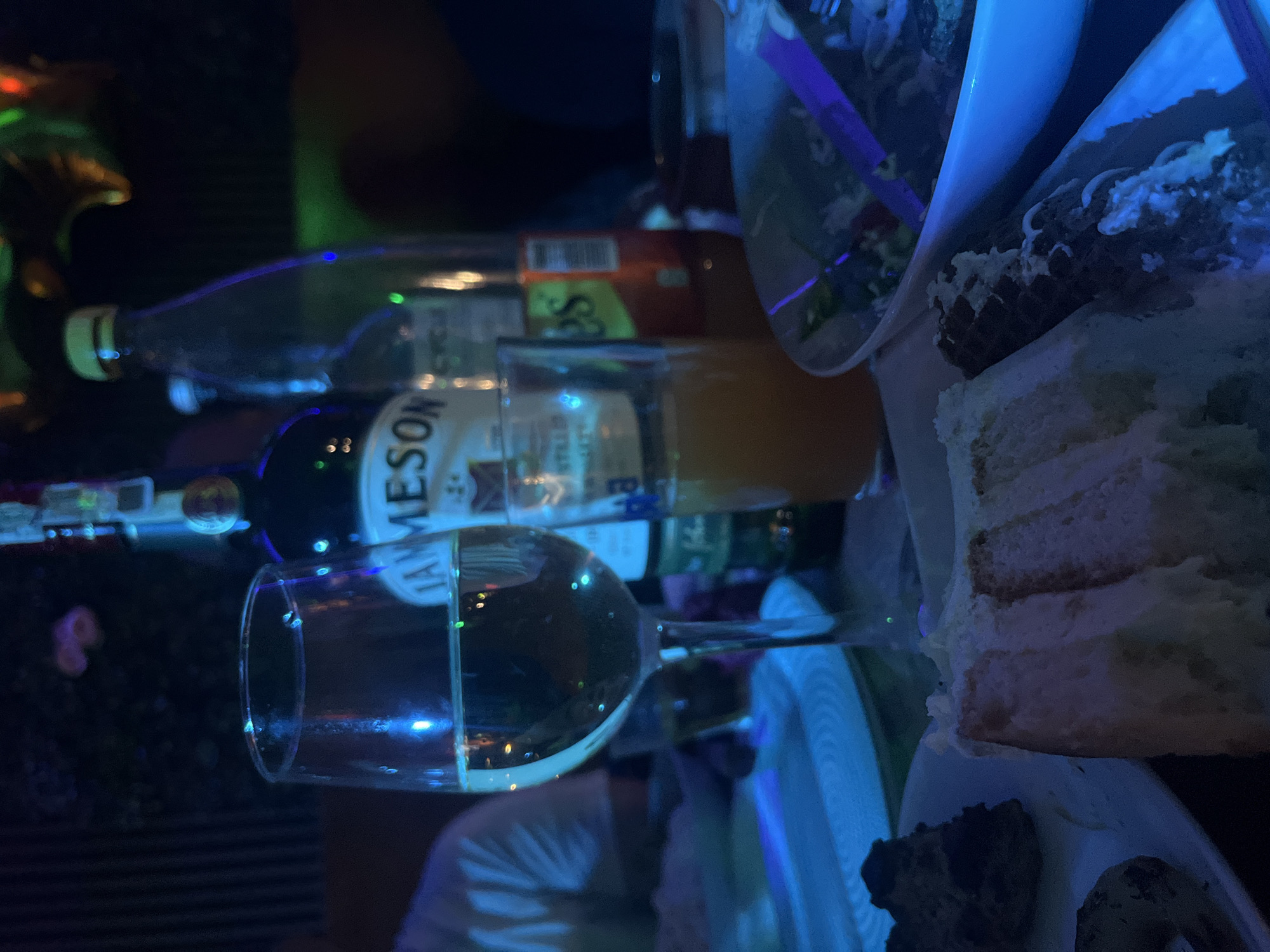 A horizontally-oriented photograph depicts a dark indoor scene, with a variety of items on a table. There is a half-full wine glass, a bottle of Jameson whiskey, another bottle of what appears to be vodka or gin, and a container of orange juice. Additionally, there are dishes on the table with a slice of cake, some sort of dark-colored food, and a salad in a container with a purple utensil inside. A person is visible in the lower center of the image, wearing a light-colored shirt. The presence of alcoholic beverages and food items suggests a gathering or celebration. No information in the picture indicates the location of the city of Lozarevo, Bulgaria.