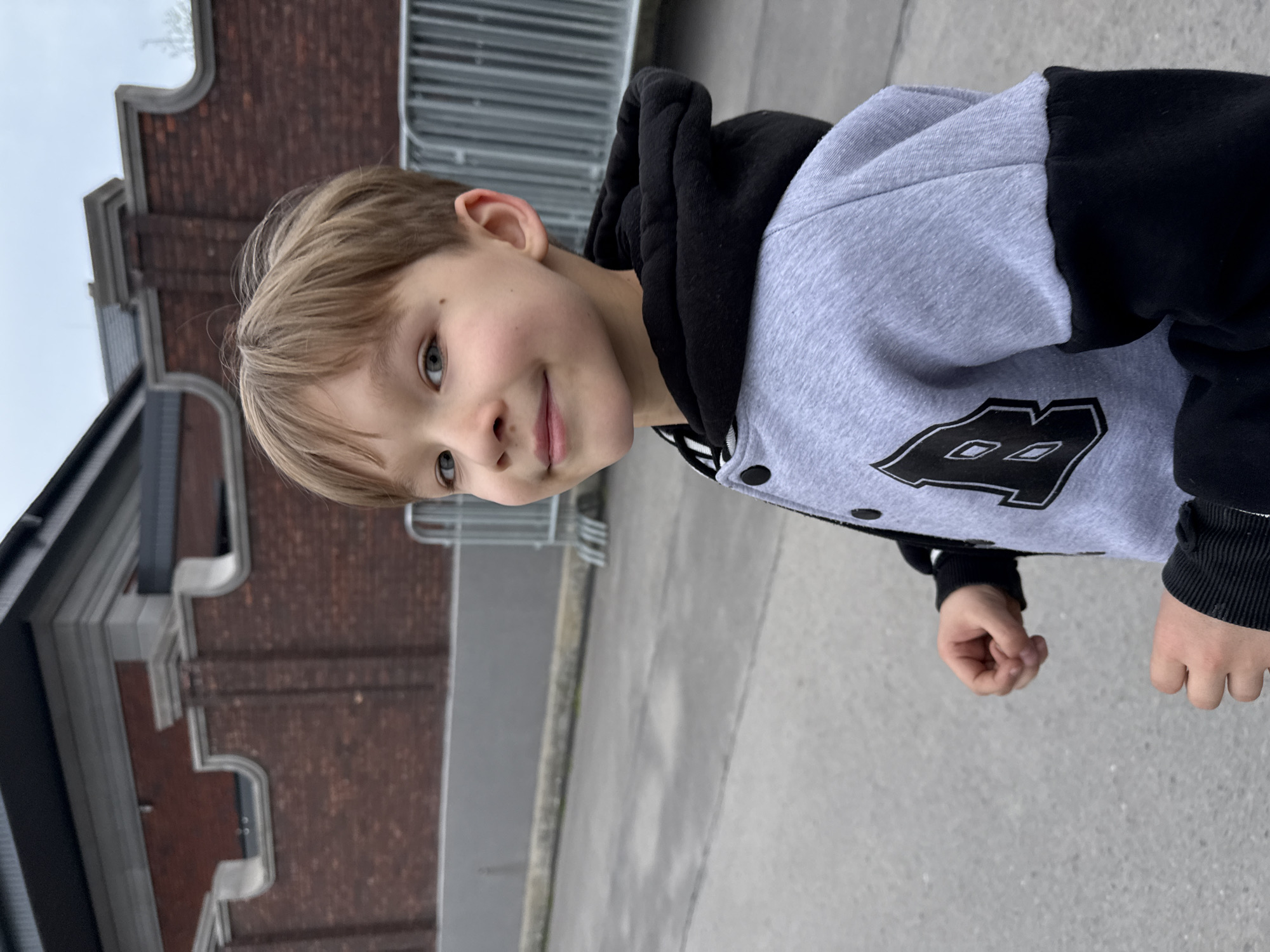 A young boy stands on a paved surface, likely a sidewalk or street. He's wearing a grey and black hooded jacket. The jacket has a prominent design on the front. Behind him are brick buildings, metal fencing, and a concrete walkway. The scene does not appear to be in Rostov-on-Don.