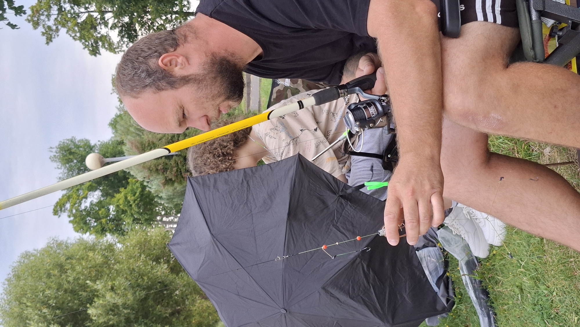 An adult male with a shaved head and beard sits in a folding chair, facing right, with a fishing rod in his hands. He is wearing a black t-shirt and black shorts with white stripes. Next to him, a child with curly hair is seated, partially obscured by a black umbrella. The child wears a light-colored t-shirt with graphics. The fishing rod is yellow and white, with a fishing reel attached. The scene appears to be a park or outdoor area with green grass and trees in the background. The sky is overcast. The image does not provide specific details to pinpoint a location within the city of Myadzyel, Belarus.