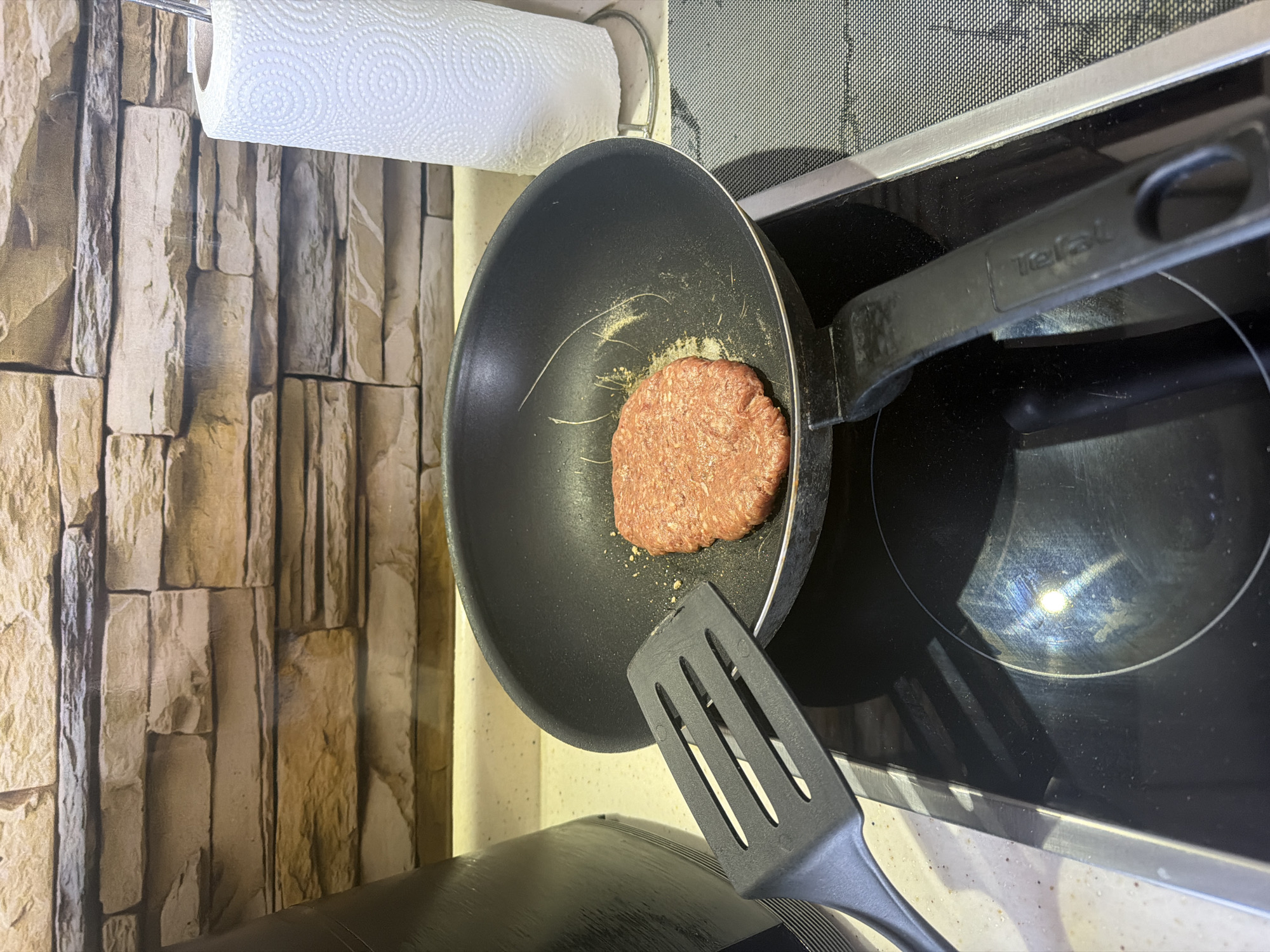 The image presents an overhead view of a kitchen counter in Shumen, Bulgaria, focusing on a stovetop and cooking preparations. A black non-stick frying pan, bearing the "Tefal" brand name on its handle, is situated on a black ceramic induction hob. Inside the pan, a single, raw, reddish-pink hamburger patty is visible, placed towards the lower right side of the pan's surface. Next to the burger, several thin, light-colored strands or hairs, along with some granular specks, are noticeable on the pan. A black, slotted plastic spatula is positioned in the lower foreground, with its head resting near the pan, suggesting it is poised for use.
To the left of the cooking area, a wall is adorned with wallpaper or tiles designed to mimic light-colored stacked stone or brick. Above this wall, a roll of white, textured paper towels is mounted on a metallic holder. The scene is brightly lit, with reflections visible on the glossy surface of the stovetop. The overall setting captures the initial moments of preparing a meal in a contemporary kitchen environment.