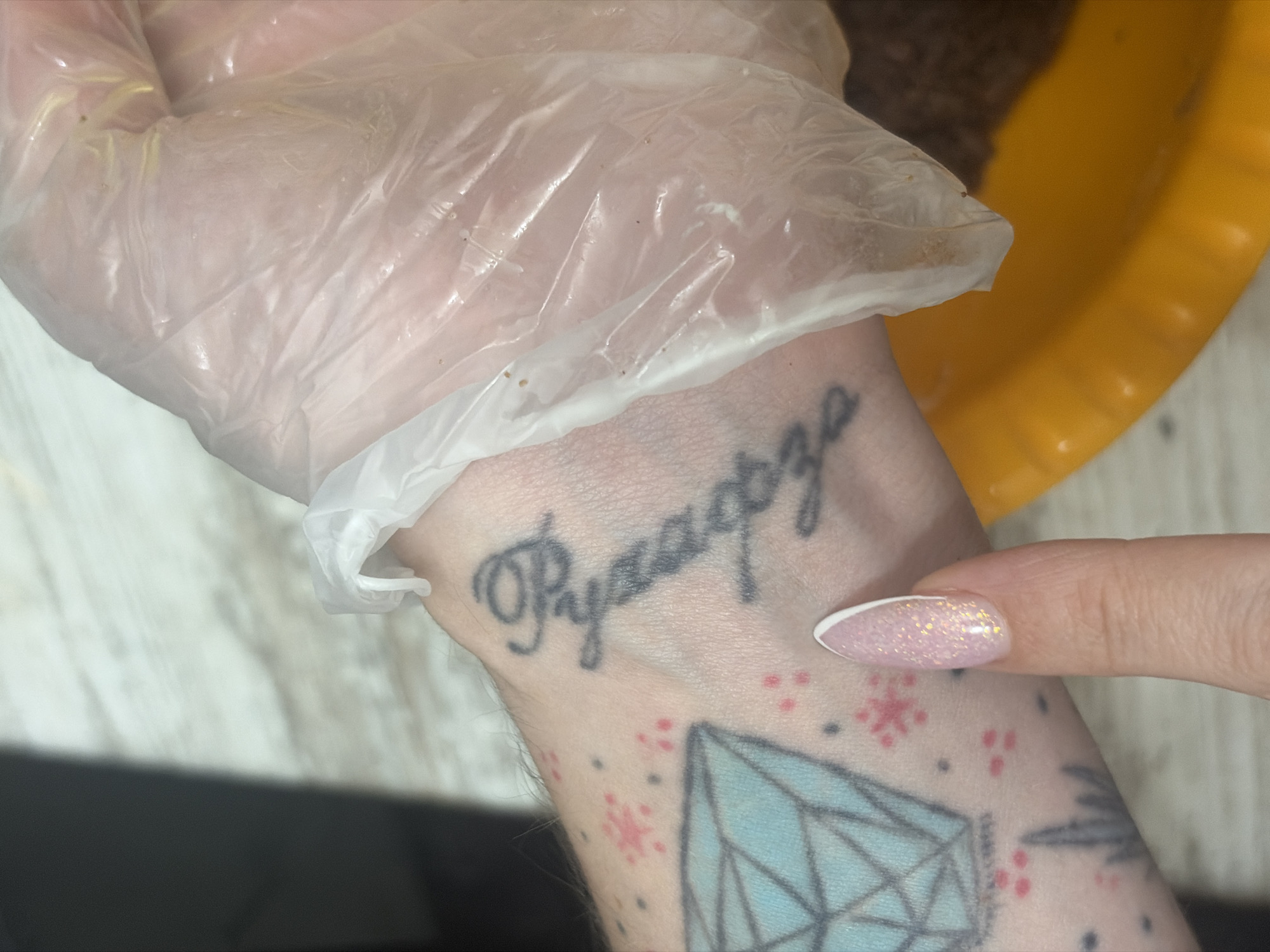 A gloved hand is seen holding another arm with tattoos. The tattoo on the wrist reads "Pyaagorza" in Cyrillic script. Below the wrist, there's a tattoo of a diamond and red and black dots. A finger with pink glitter nail polish points to the wrist. A yellow container is in the upper right corner. The background is a light-colored surface. The Cyrillic script tattoo likely says "Петербургa", which means "of Saint Petersburg". Thus, the tattoo represents the location of the city of Saint Petersburg, Russia.
