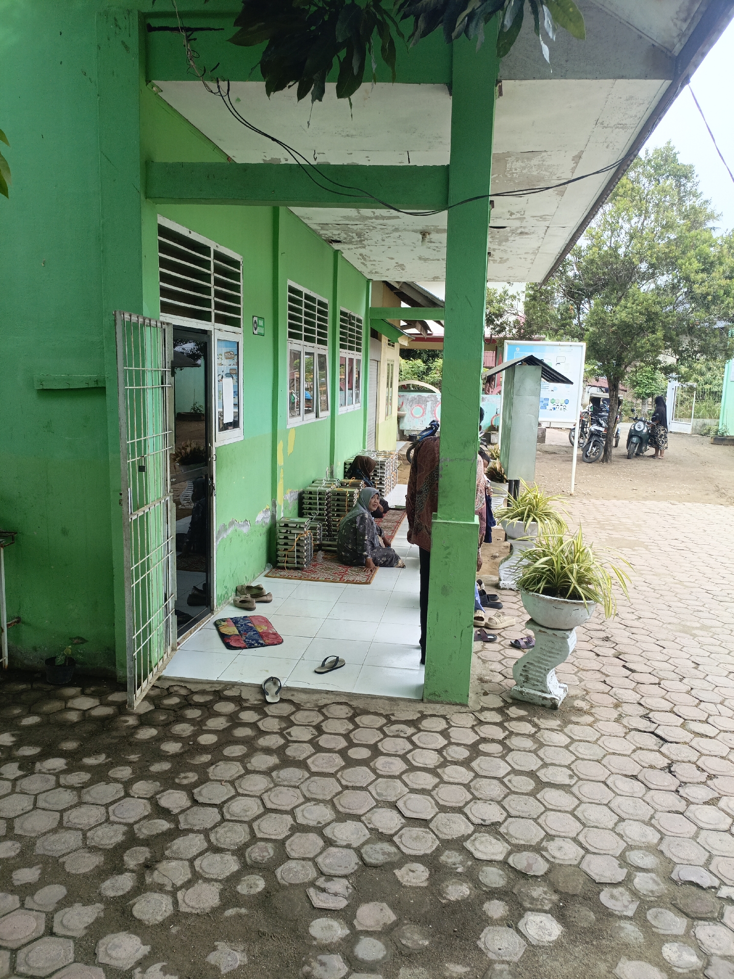 The image depicts the exterior of a green building, likely a school or community center, with a covered walkway. Several people are present, some sitting on mats in front of the building, others standing near motorcycles in the background. The ground is paved with hexagonal bricks and soil. A few potted plants add greenery to the scene. In the background, there is a notice board and trees. The setting appears to be a public space in Lhokseumawe, Indonesia.