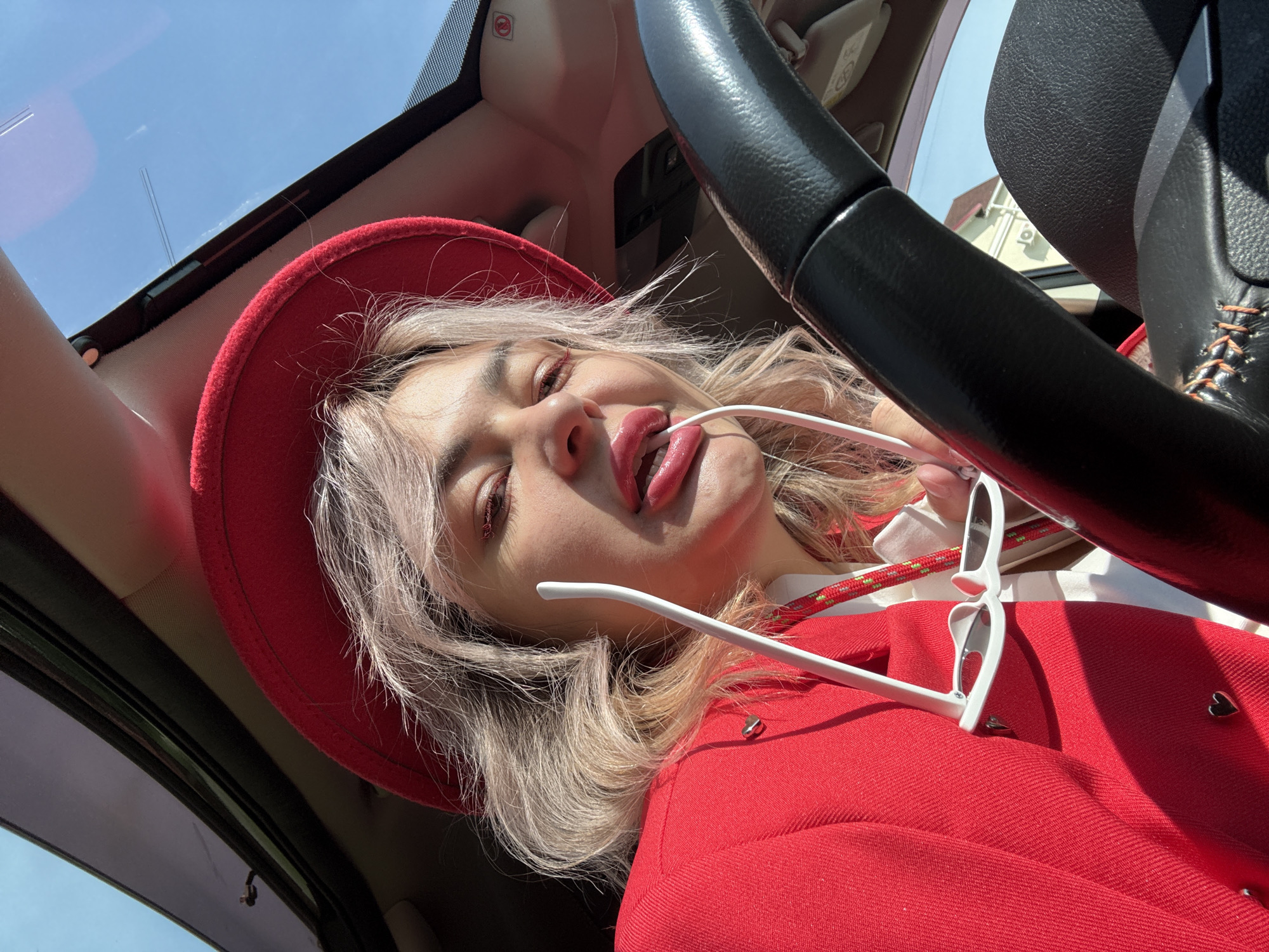 A woman wearing a red hat and red blazer is in the driver's seat of a car, looking at the camera. She is holding white sunglasses in her mouth and has bright pink lipstick on. The woman's hair is blonde and wavy. The car's sunroof is open, revealing a blue sky. The steering wheel of the car is visible in the foreground. The scene is lit by sunlight. The location is not specified in Birobidzhan, Russia.