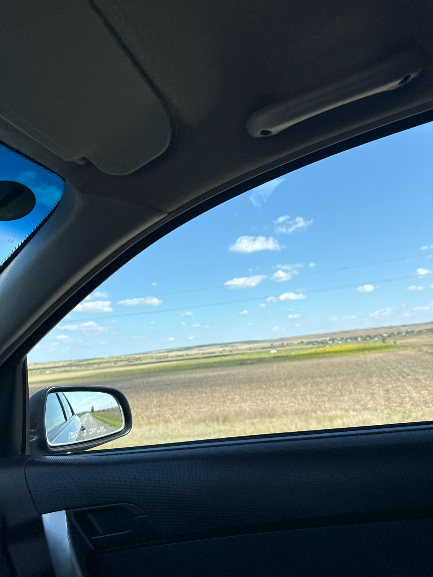 The image shows a view from inside a car looking out the window and side mirror. The sky is blue with scattered white clouds. Beyond the window, there is a flat landscape with fields of dry grass and some green vegetation in the distance. A road is visible in the side mirror, with power lines stretching across the horizon. The interior of the car, including the sun visor, door panel, and a handle, is also visible. The location is specified as Carabetovca, Moldova.