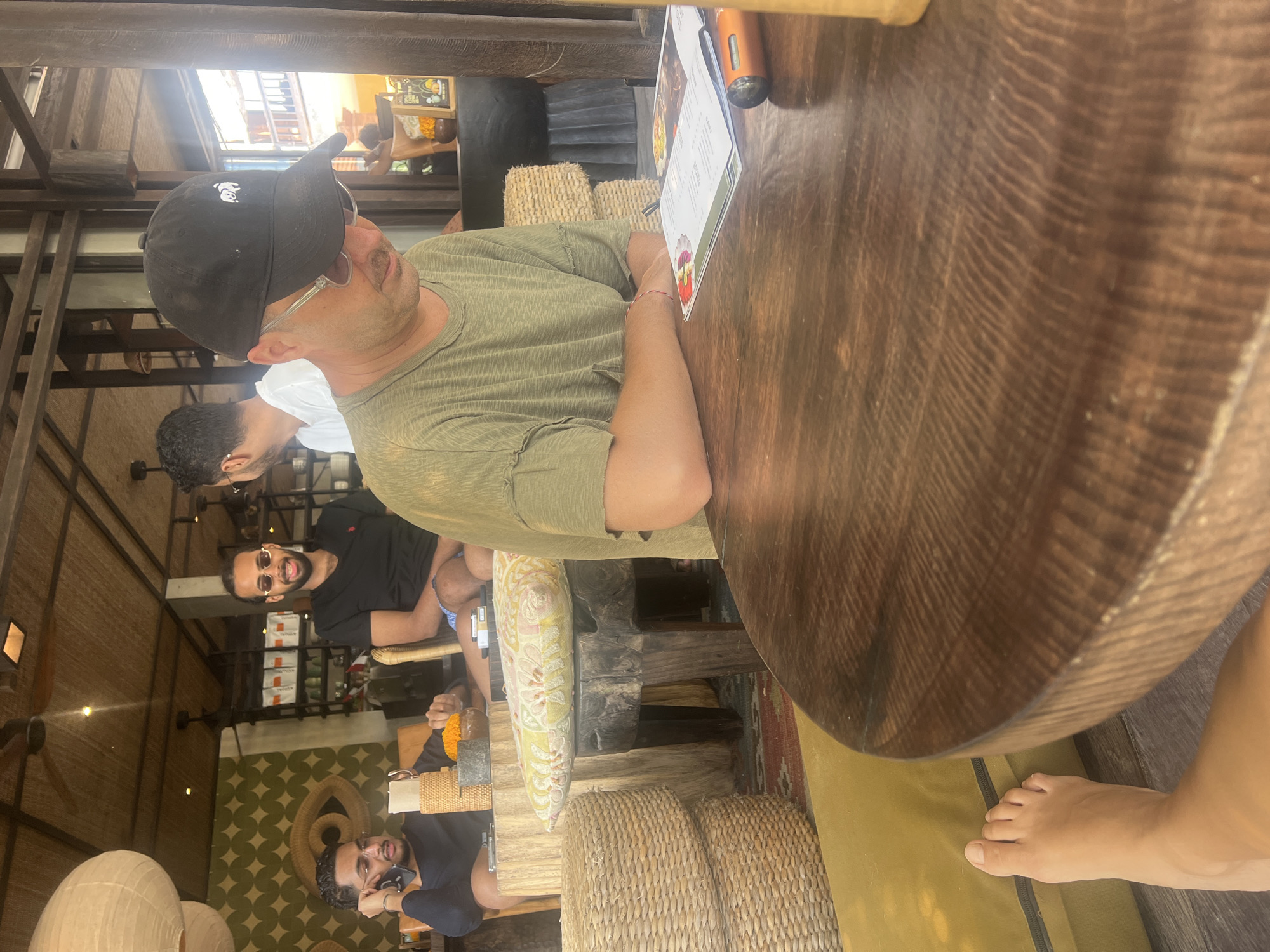 The image depicts an indoor dining scene in Mengwi, Indonesia. In the foreground, a person wearing a green t-shirt and a black baseball cap with a white logo is seated at a wooden table, looking down at a menu and an orange object. To the left, another person is on the phone, and a third person is visible in the background wearing a black t-shirt and sunglasses. The environment appears to be a restaurant or cafe with thatched roofing, woven chairs, and decorative elements like a large eye-shaped tapestry on the wall. Sunlight filters into the space, creating a warm ambiance. A bare foot is visible in the lower left corner of the frame, resting on a yellow cushion.