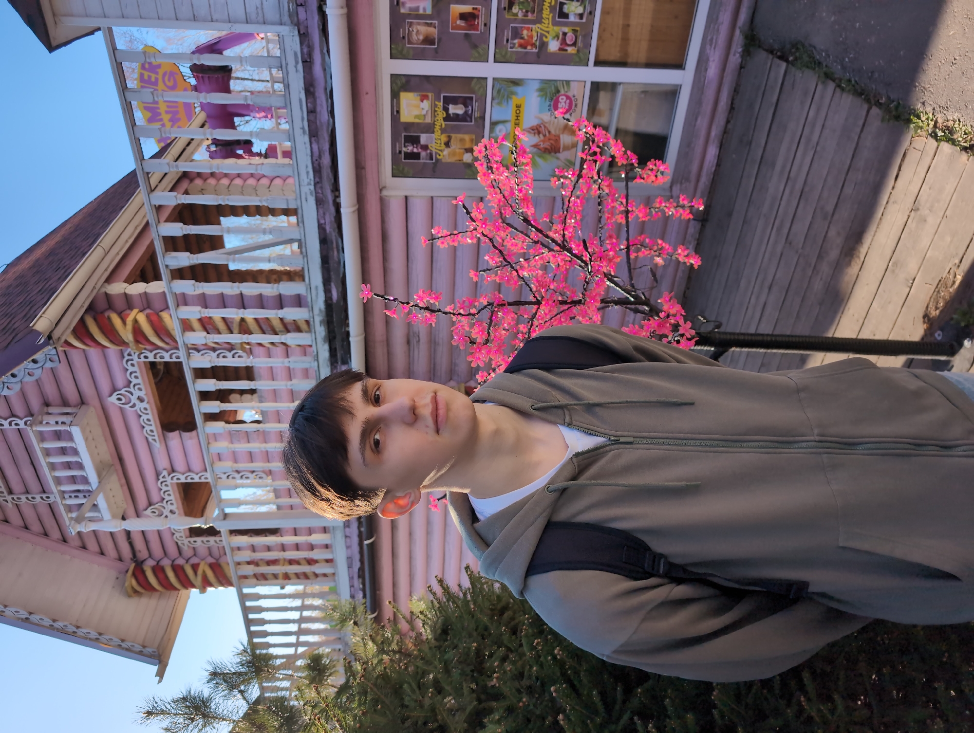 A person wearing a green hoodie and a backpack stands in front of a light pink building in Izhevsk, Russia. The building has white trim and a sign that says "MEVER WIN". A pink tree stands to the right of the person. The ground is paved with wood planks. Green shrubbery is visible in the foreground. The sky is blue.