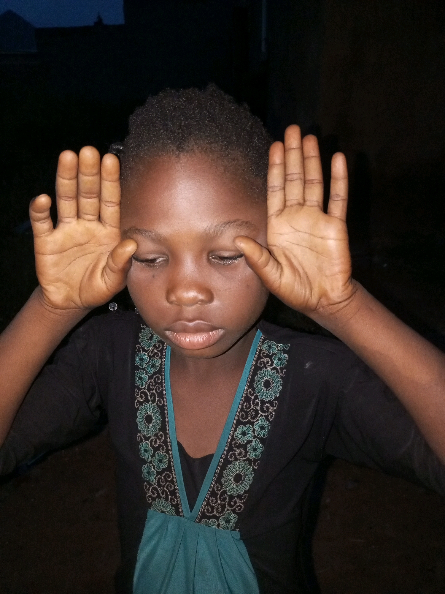 In a dimly lit outdoor environment, a young person with dark skin and short, textured hair is shown from the chest up, looking downwards with closed eyes and hands held up near their temples. The person is wearing a dark shirt with intricate teal and black embroidery around the neckline and a teal fabric panel below the embroidery. The background is dark and indistinct, suggesting nighttime or a poorly lit area, with faint outlines of structures visible in the upper left corner. The overall lighting suggests this was taken with a flash, illuminating the subject's face and hands against the dark surroundings. The scene appears to be in a residential area within Tempure, Nigeria.