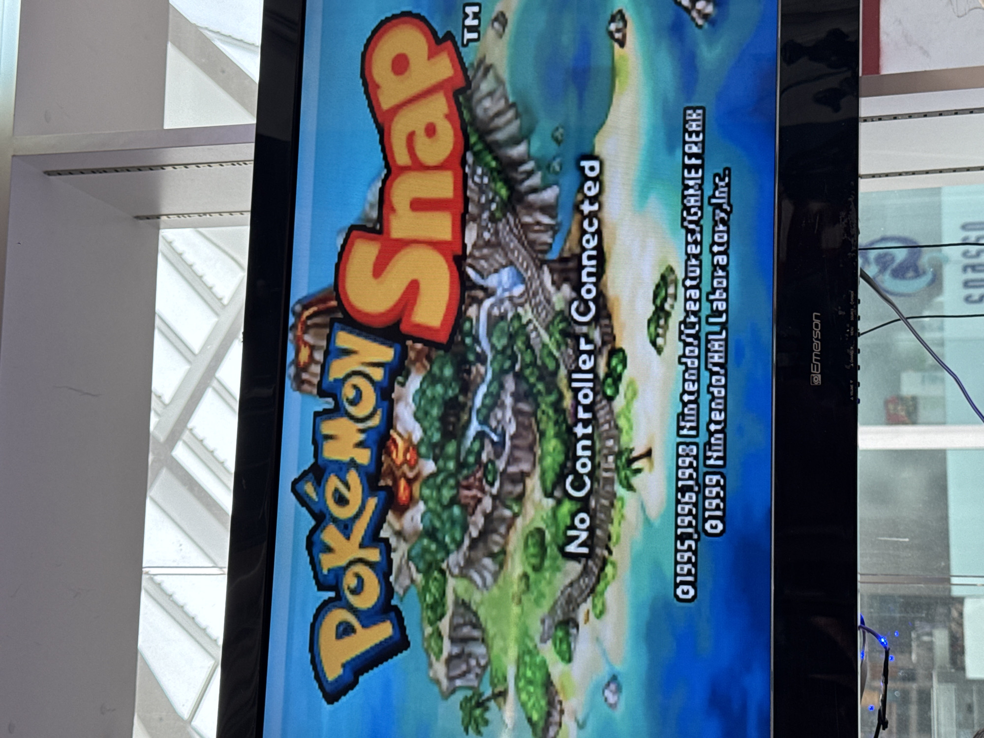 An image displays the title screen of the video game "Pokémon Snap" on a television. The game's title, "Pokémon Snap," is prominently featured in large, colorful text across the top portion of the screen. Below the title, a vibrant, tropical island landscape is depicted, complete with lush greenery, sandy beaches, and clear blue water. A winding path is visible on the island, suggesting a gameplay element. Centered on the screen, in white text, is the message "No Controller Connected," indicating the game is paused or awaiting input. At the bottom of the screen, copyright information is visible, listing "©1995, 1996, 1998 Nintendo/Creatures/GAMEFREAK" and "©1999 Nintendo/HAL Laboratory, Inc." The television itself is black and appears to be an older model, with visible buttons and branding on its lower bezel that reads "Emerson." The television is positioned in front of a window that allows natural light to enter, illuminating the scene. Through the window, the ceiling structure of what appears to be a commercial space, possibly a store or exhibition hall, is visible. The image seems to have been taken in El Paso, United States of America.