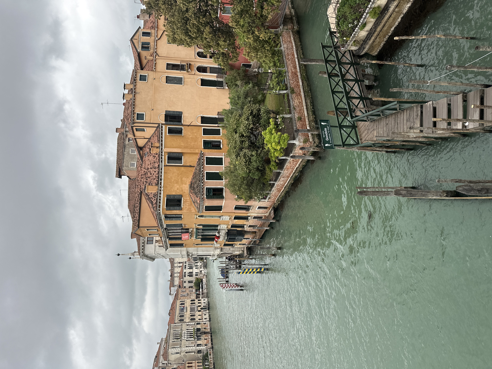 The image depicts a canal in Venice, Italy, under a cloudy sky. On the left, multi-story buildings with orange facades and tiled roofs line the canal. Windows, many with closed shutters, punctuate the buildings. A small canal-side garden with greenery sits adjacent to one building. A wooden bridge with green metal supports crosses the canal to the right, leading to a pathway. The water in the canal is a murky green color. Warning pylons mark the water. Buildings are visible in the distance on the left.